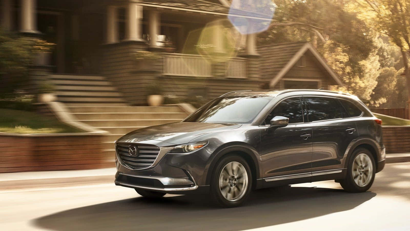 Mazda Cx-9 桌布 壁紙 — 一輛時尚的灰色SUV駛過陽光明媚的郊區房屋，背景帶有動態模糊效果。