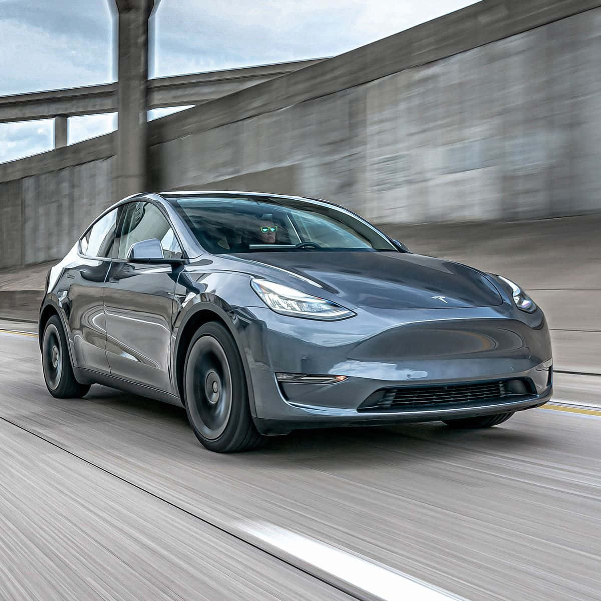 特斯拉 桌布 壁紙 — 一輛時尚的灰色Tesla Model Y在混凝土高速公路上行駛，經過天橋下方，動態模糊強調了速度。