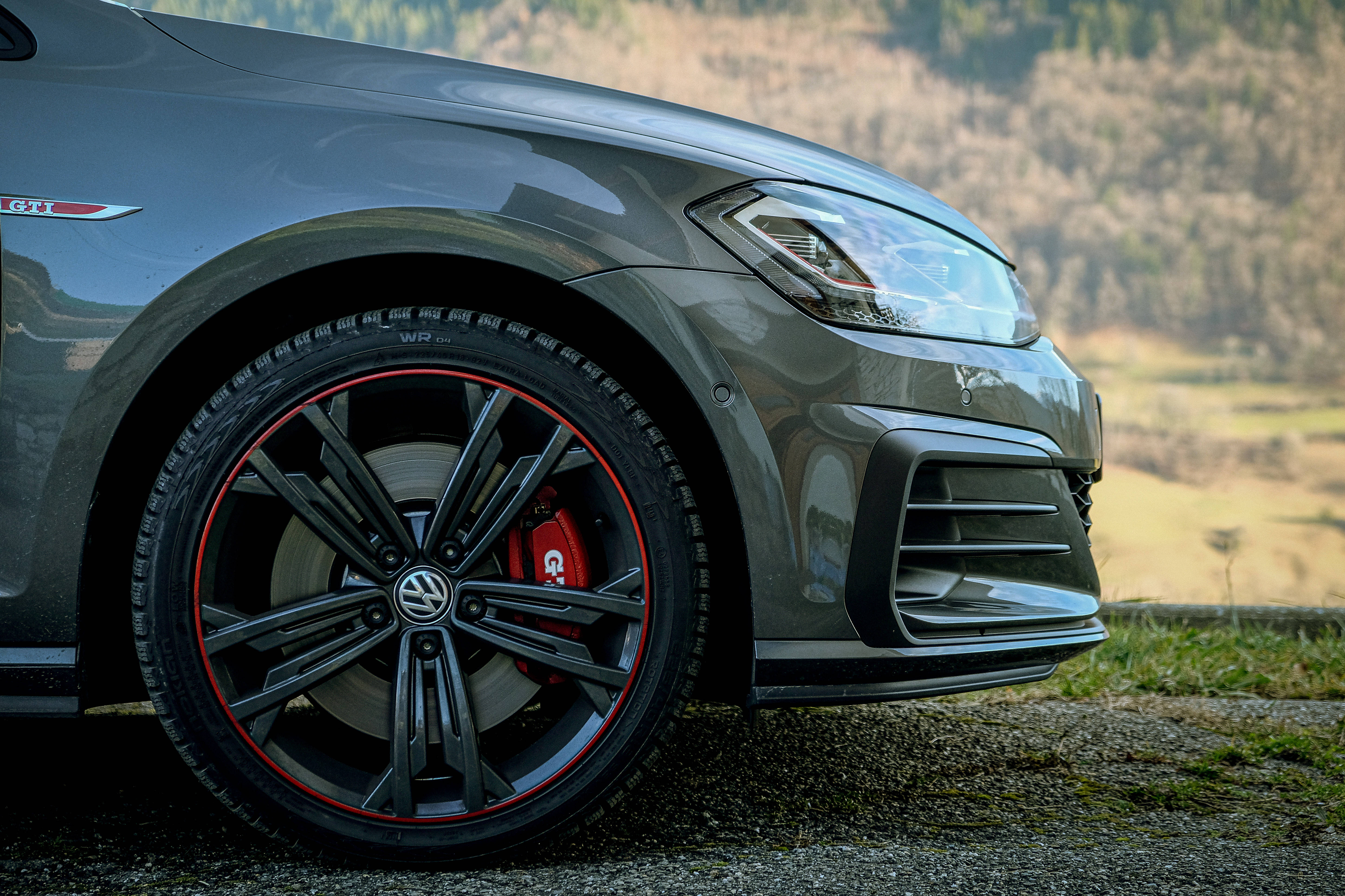 Gti 桌布 壁紙 — 一輛時尚的灰色Volkswagen GTI，配備紅色制動卡鉗和黑色輪圈，停泊在草地山坡旁的碎石路上。