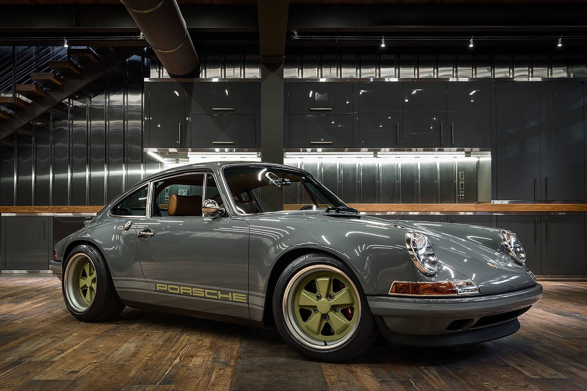 Singer 桌布 壁紙 — 一輛時尚的灰色保時捷911配金色輪圈，在現代工業車庫的攝影棚燈光下閃閃發光，車庫內有深色櫥櫃和木地板。