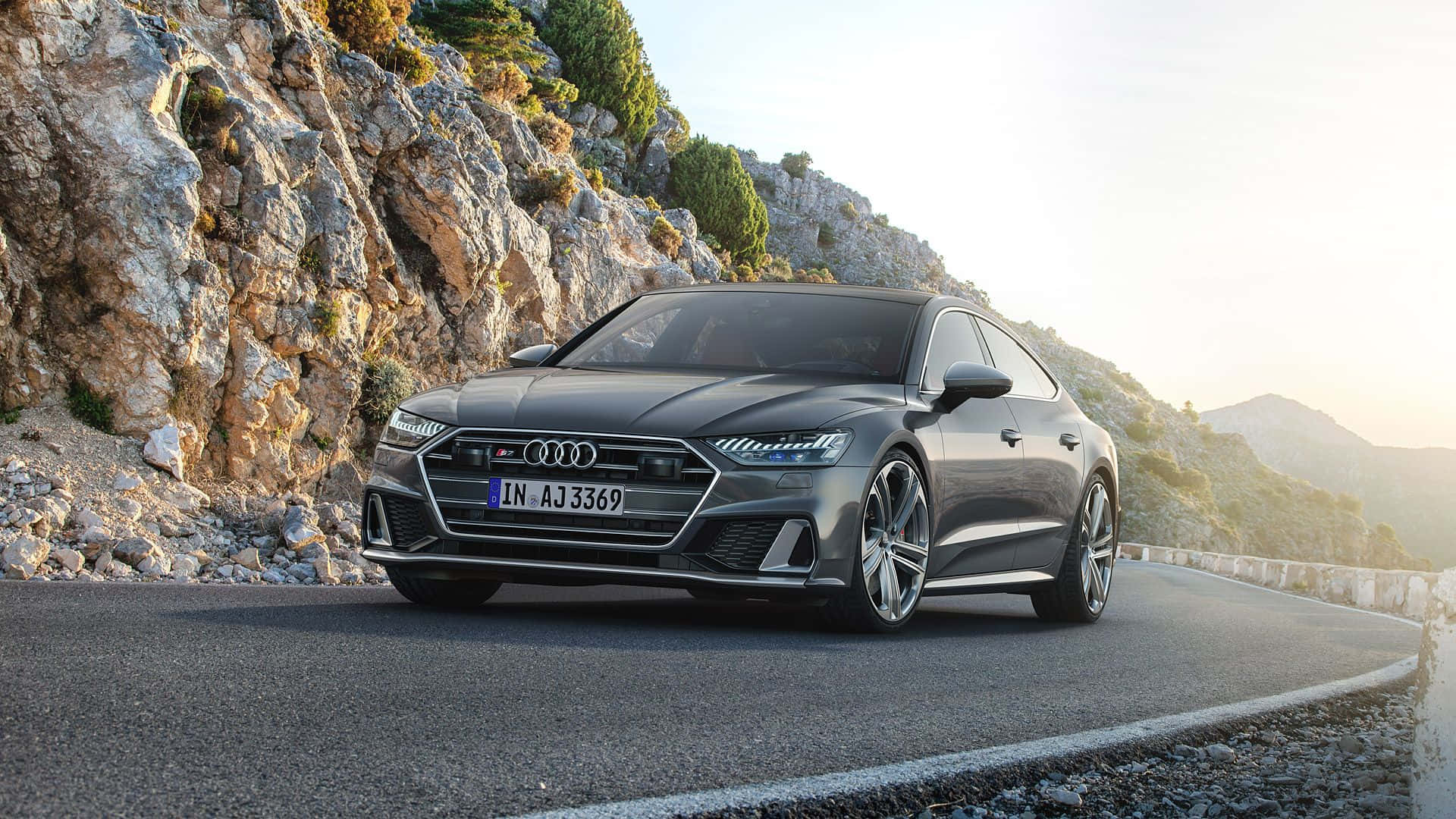 Audi S7 桌布 壁紙 — 一輛時尚的灰色奧迪 RS7 Sportback 在黃金時段沿著蜿蜒的山路巡航，崎嶇的懸崖和遠處的山丘沐浴在溫暖的陽光下。