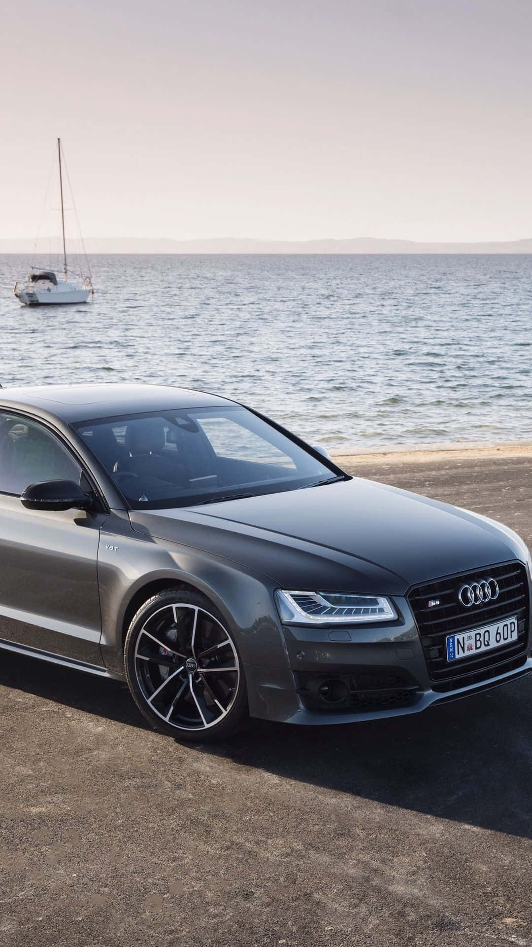 Audi S8 桌布 壁紙 — 一輛時尚的灰色奧迪S8停在海邊，一艘帆船在平靜的水面上漂浮，天空朦朧。