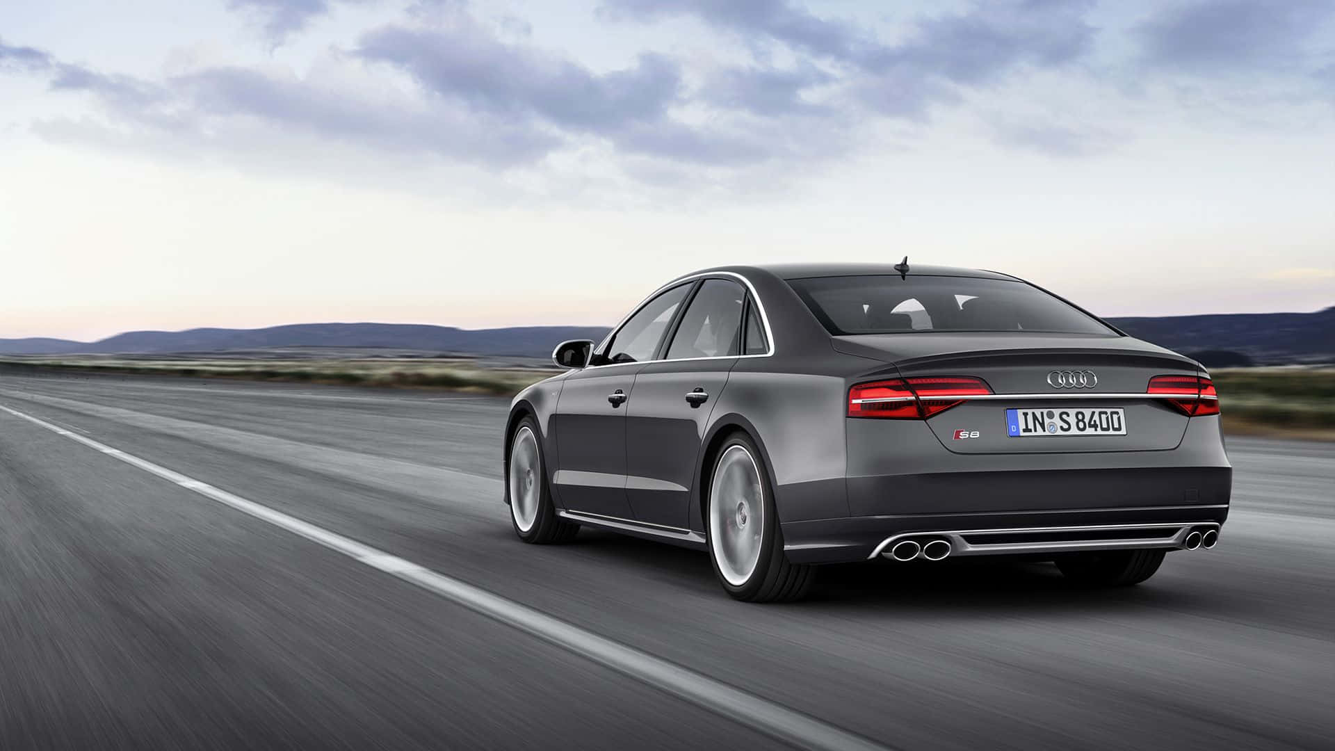 Audi S8 桌布 壁紙 — 一輛時尚的灰色奧迪S8轎車在黃昏時分，於多雲的天空下，沿著一條漫長開闊的道路疾馳，呈現模糊的動態效果，遠方山丘隱約可見。