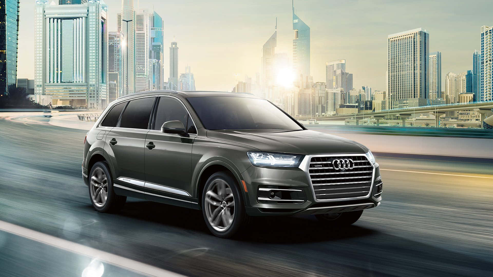 Audi Q7 桌布 壁紙 — 一輛時尚的灰色奧迪SUV在高速公路上奔馳，背後是現代城市天際線在金色夕陽中閃耀。
