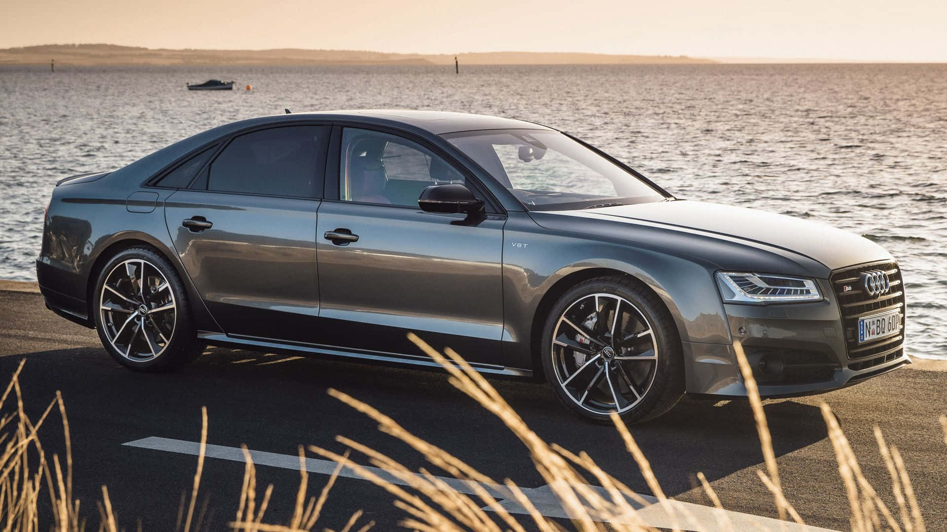Audi S8 桌布 壁紙 — 一輛時尚的灰色奧迪轎車停泊在平靜的海邊，正值黃金時刻，前景有高高的草。