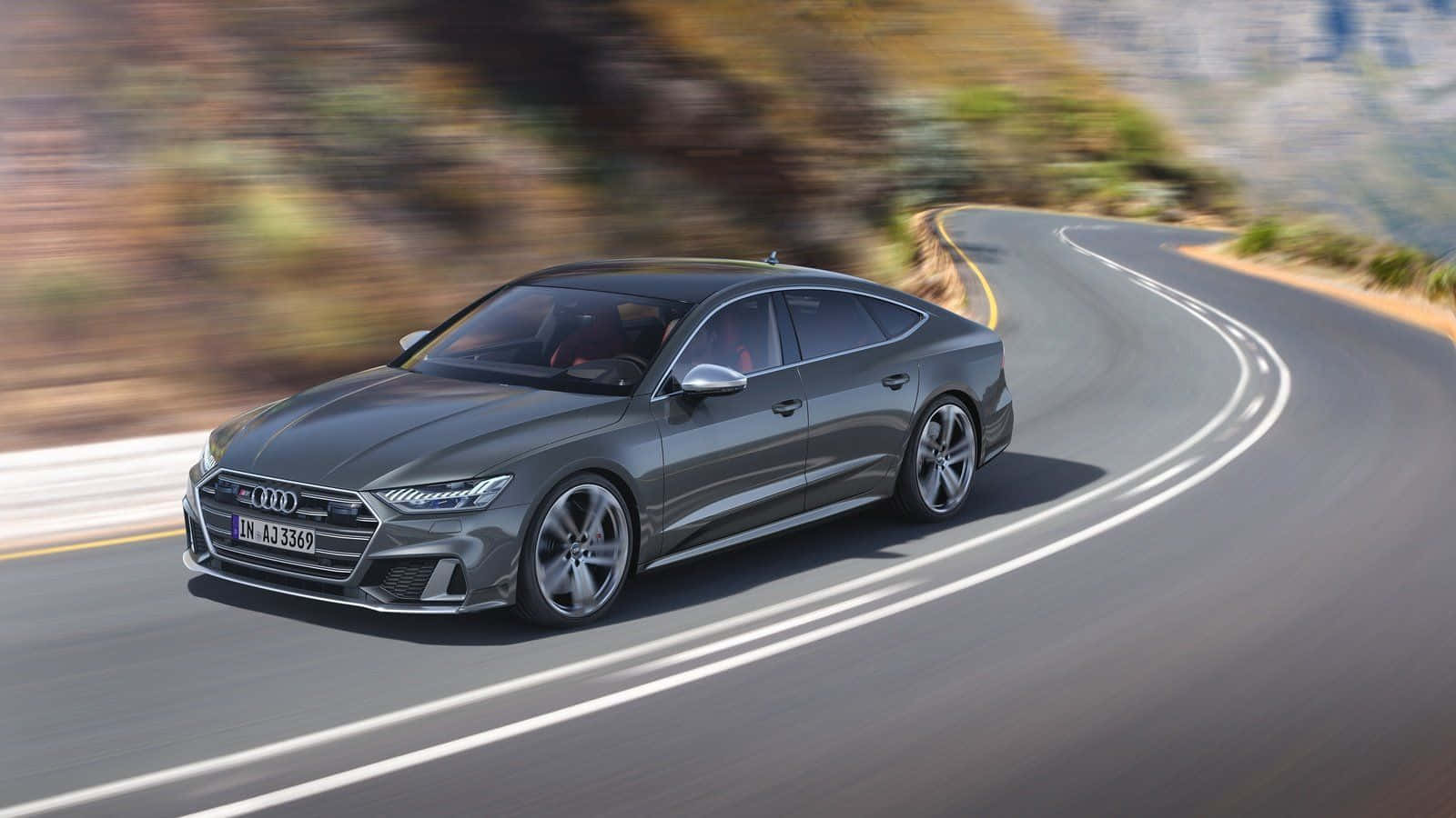 Audi S7 桌布 壁紙 — 一輛時尚的灰色奧迪轎車沿著蜿蜒的山路疾馳，背景是模糊的樹葉和遠處的山峰。