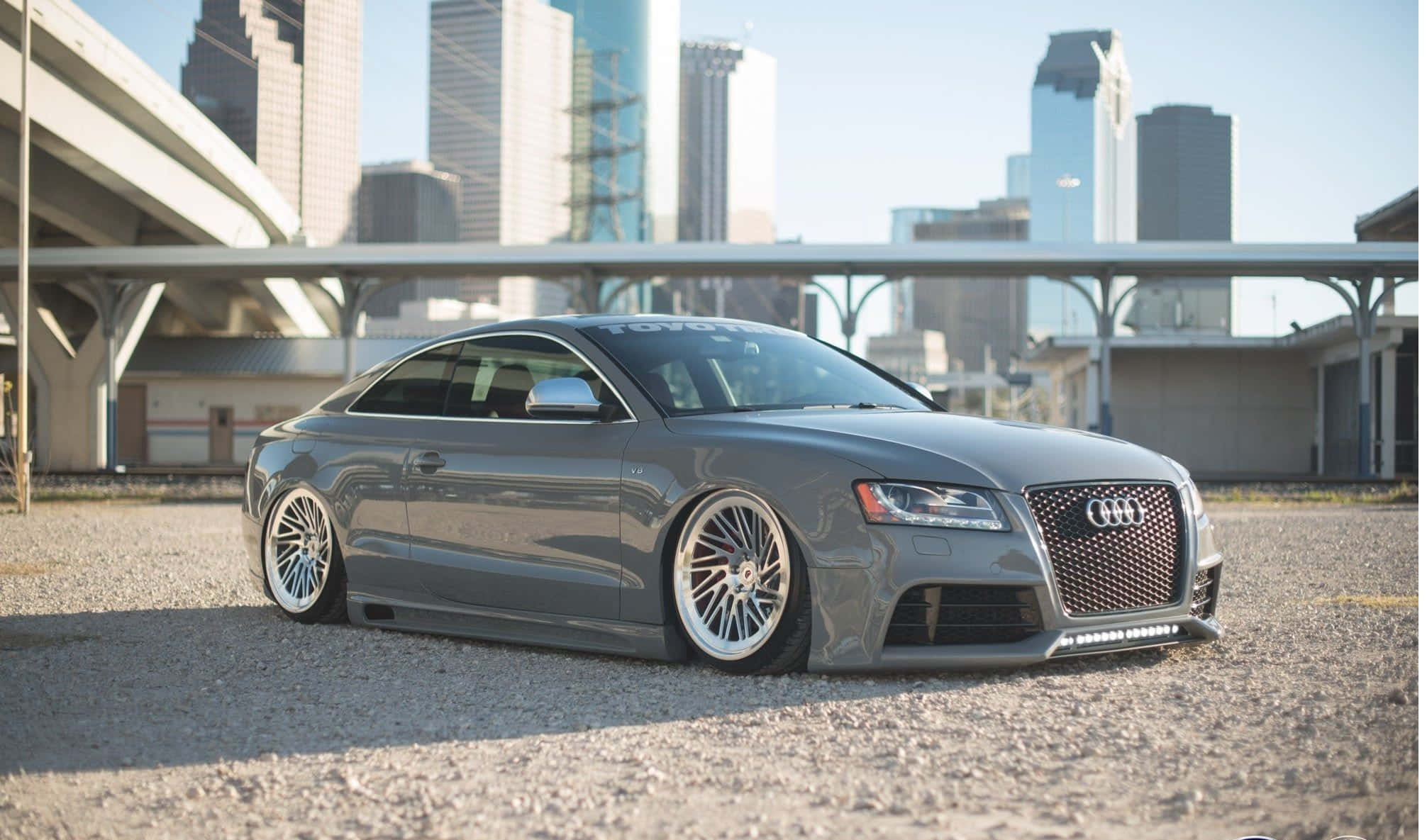 Audi S6 桌布 壁紙 — 一輛時尚的灰色奧迪雙門跑車配備定制輪圈，停泊在城市高架橋下的碎石路上，背景是高樓大廈。