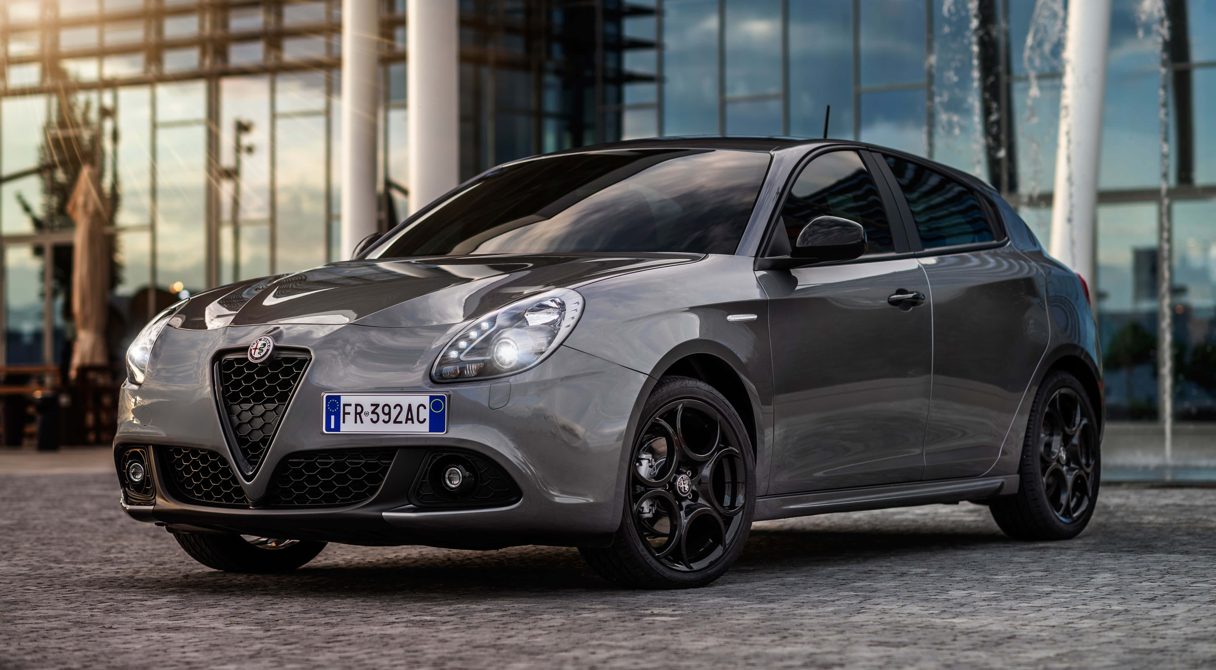 Alfa Romeo Giulietta 桌布 壁紙 — 一輛時尚的灰色愛快羅密歐Giulietta停在鵝卵石路上，前方是帶有噴泉的現代玻璃建築。