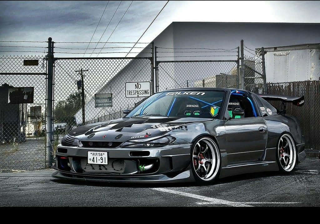 Tuned Car 桌布 壁紙 — 一輛時尚的灰色日產S14漂移車，配備客製化寬體套件和紅色制動卡鉗，停泊在陰天下的鐵絲網圍欄前。