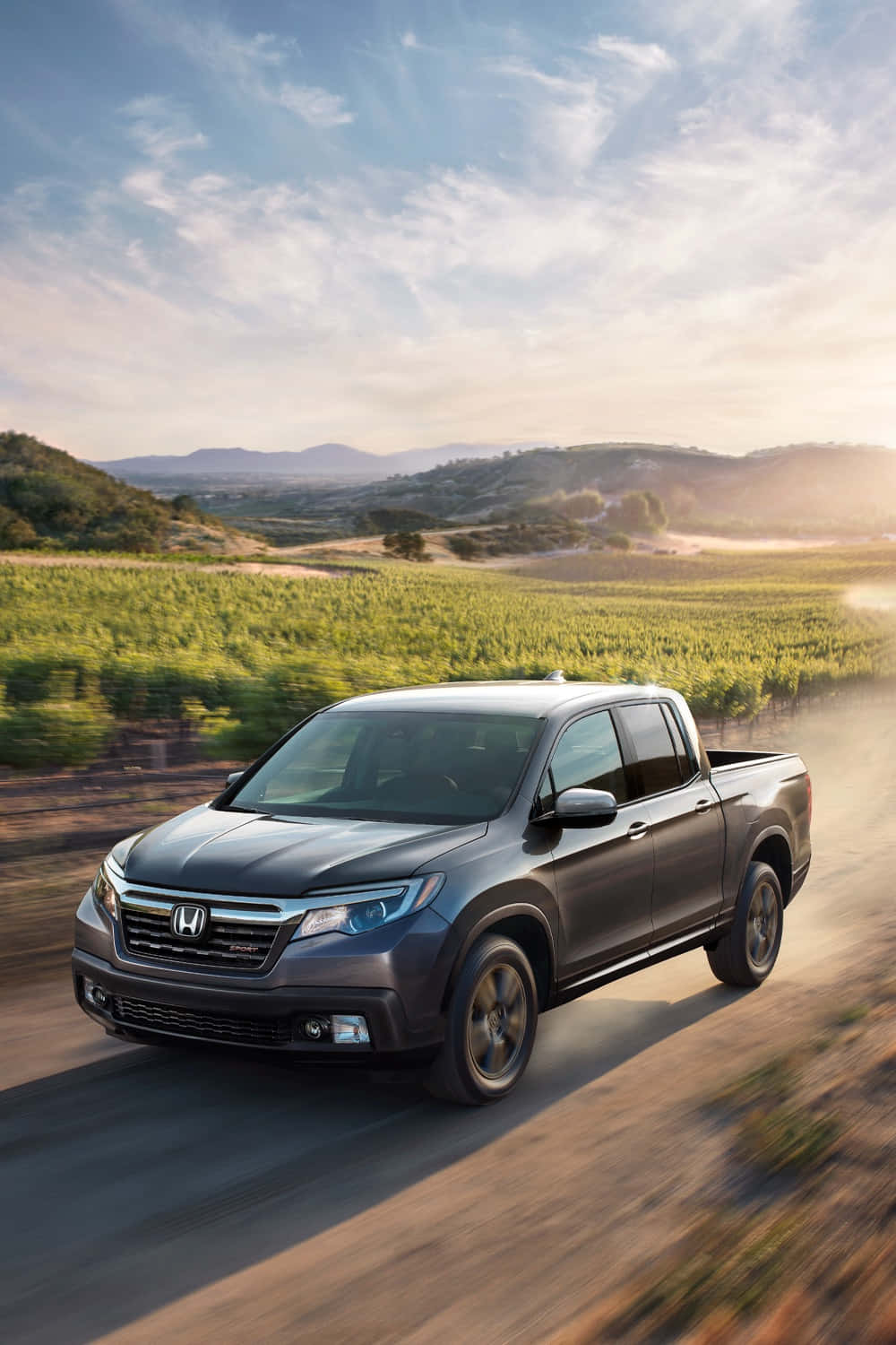Honda Ridgeline 桌布 壁紙 — 一輛時尚的灰色本田Ridgeline皮卡車在塵土飛揚的鄉間道路上疾馳，穿過陽光明媚的葡萄園，背景是起伏的丘陵。