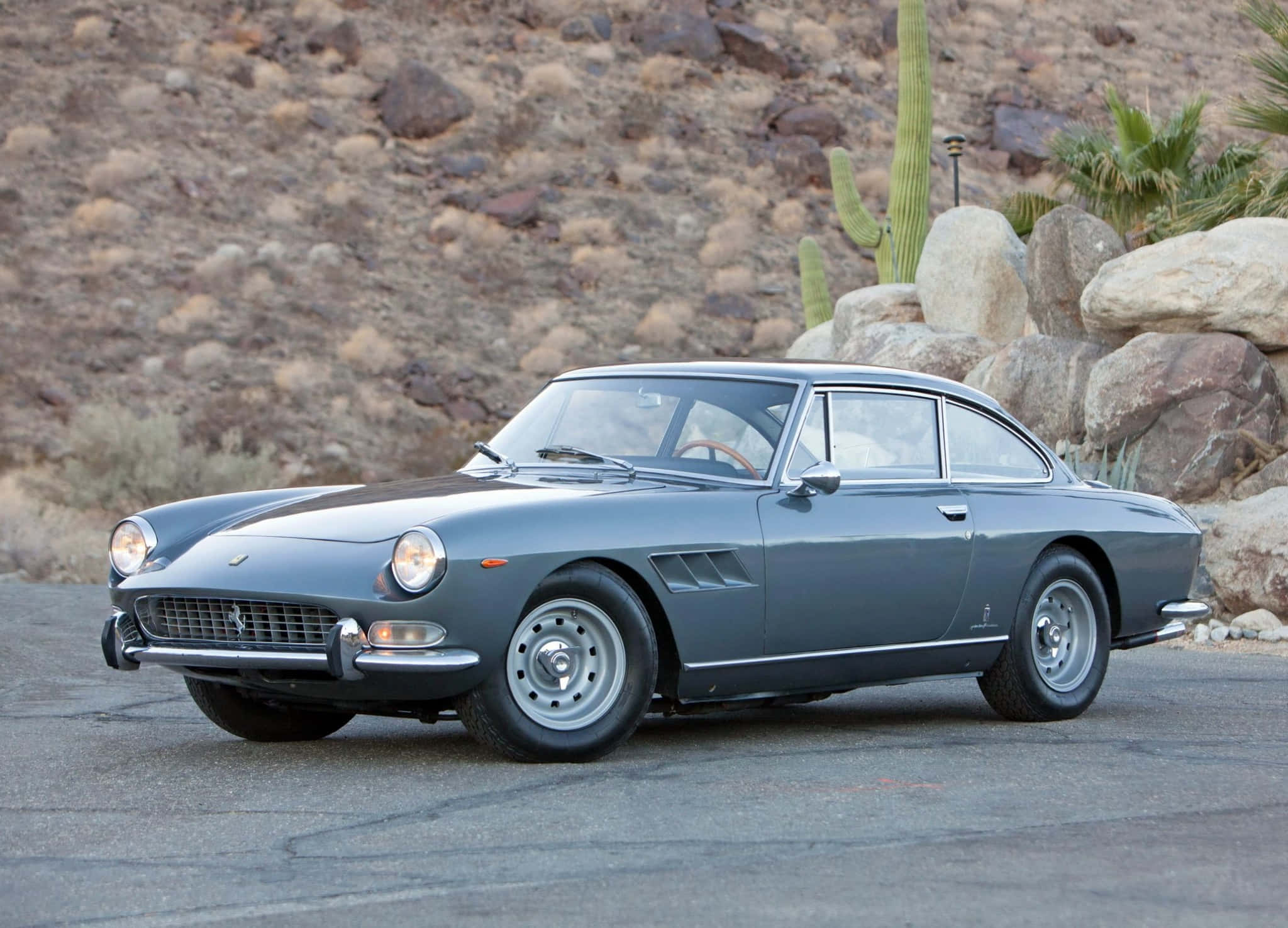 Vintage Ferrari 桌布 壁紙 — 一輛時尚的灰色法拉利275 GTB停在柏油路上，背景有沙漠岩石、仙人掌和棕櫚樹。