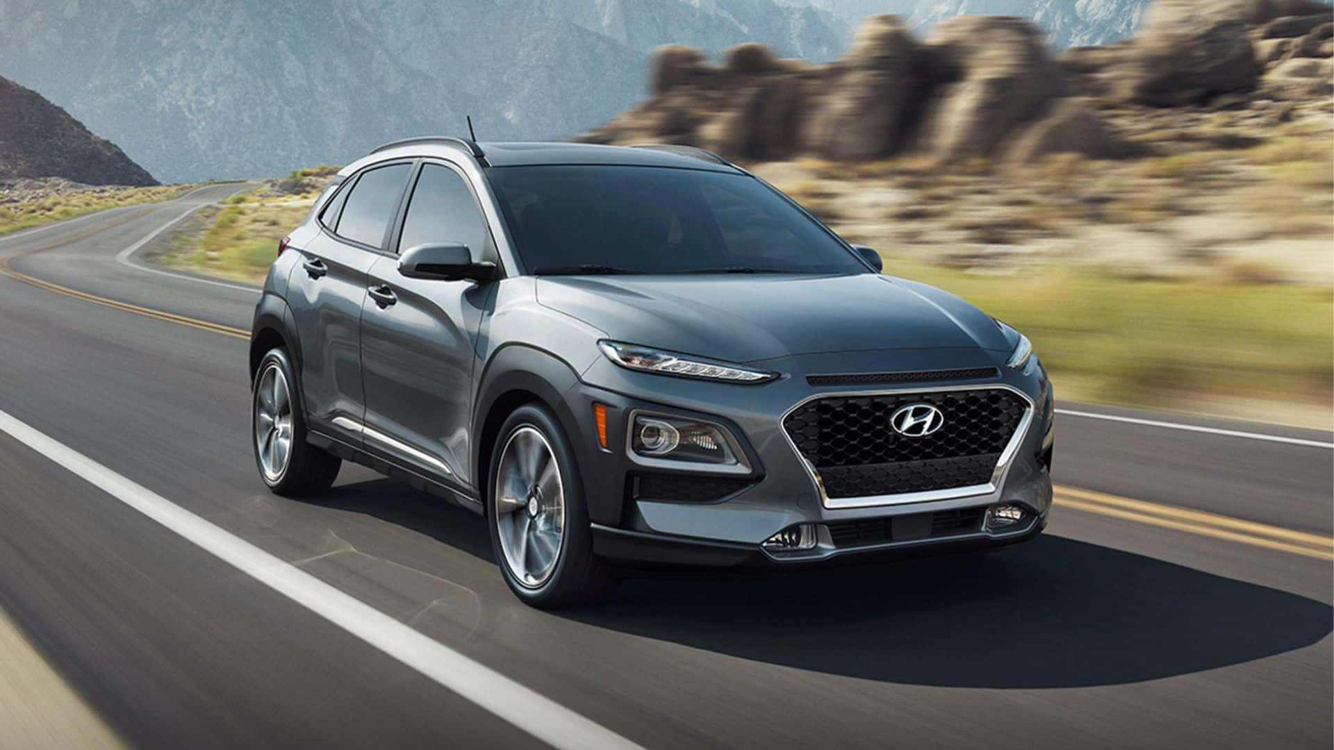 Hyundai Kona 桌布 壁紙 — 一輛時尚的灰色現代Kona SUV在蜿蜒的山路上疾馳，背景是模糊的岩石地形。