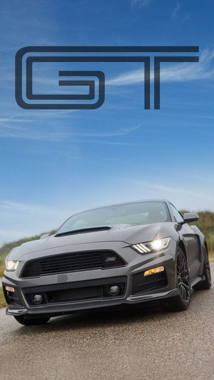 ford iPhone 桌布 壁紙 — 一輛時尚的灰色福特 Mustang GT 跑車，帶有發光的頭燈，停在瀝青路上，上方是晴朗的藍天，並有一個大型 GT 標誌。