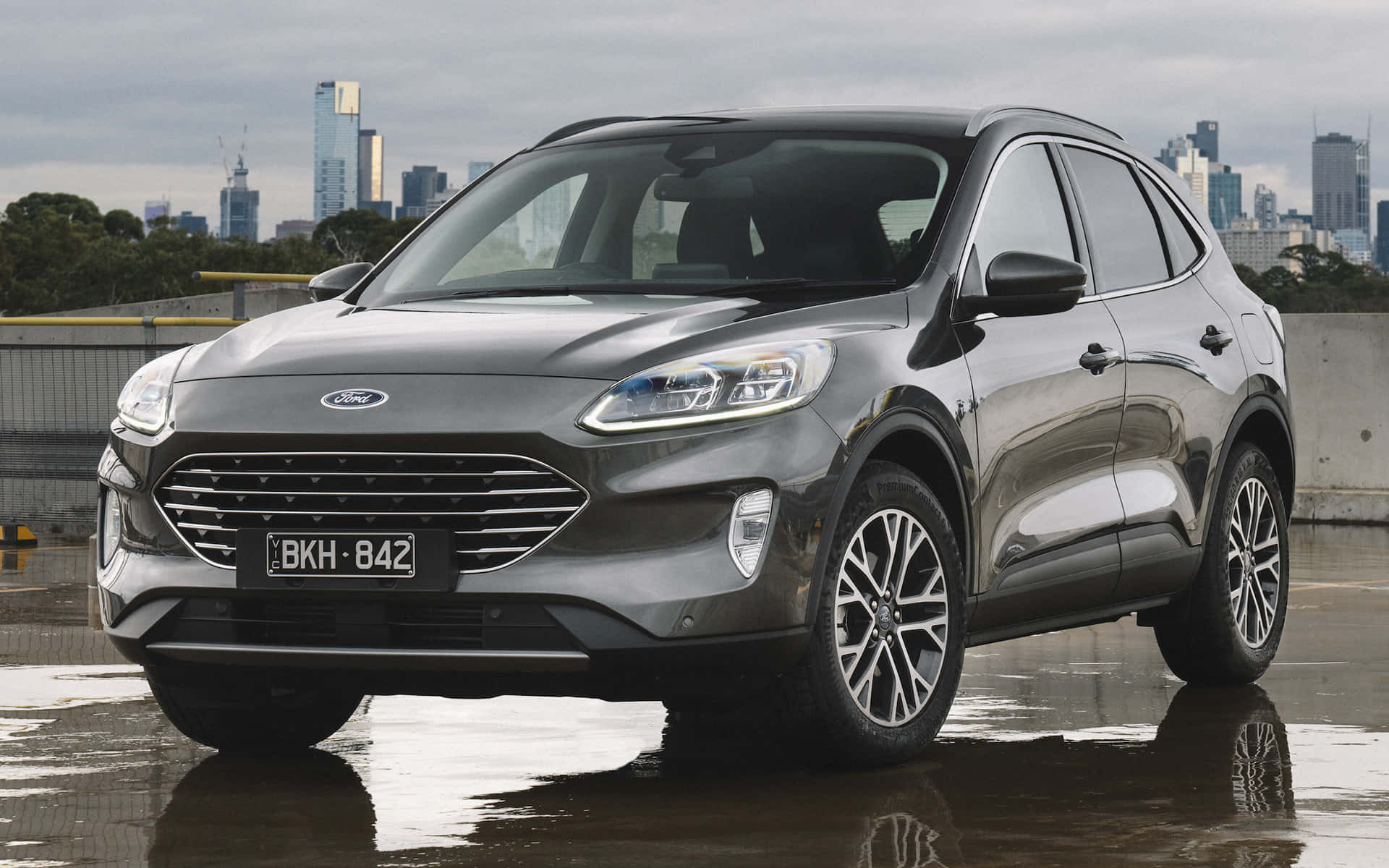 Ford Escape 桌布 壁紙 — 一輛時尚的灰色福特SUV在陰天底下反射濕漉漉的路面，背景是現代化的城市天際線。