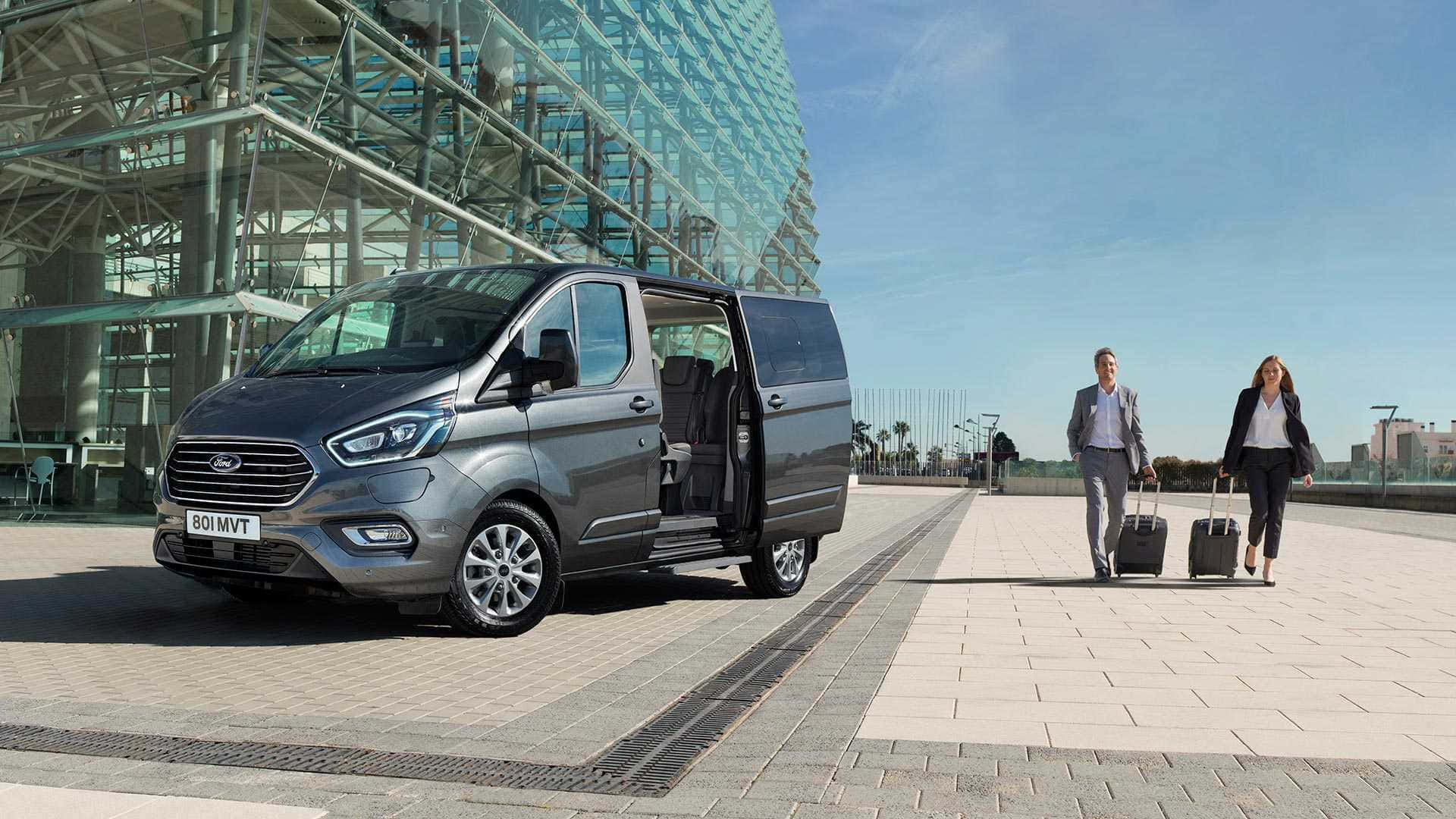 Ford Transit 桌布 壁紙 — 一輛時尚的灰色福特客貨車停放在現代玻璃建築外，兩位商務旅客在陽光明媚的日子裡拖著行李箱走過。