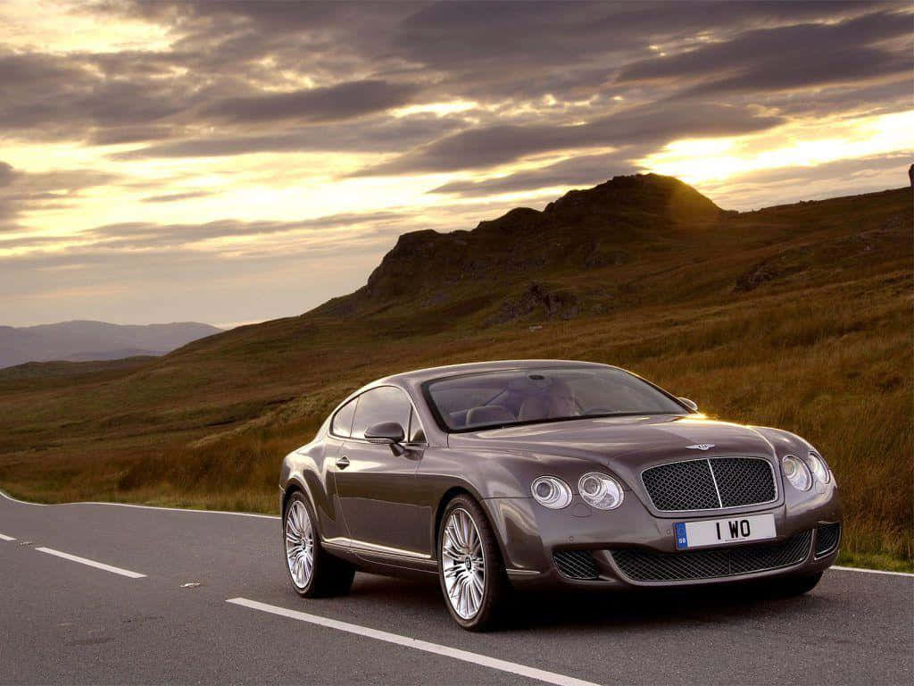 豪華車 桌布 壁紙 — 一輛時尚的灰色賓利 Continental GT 在戲劇性的金色日落天空下沿著蜿蜒的山路巡航。