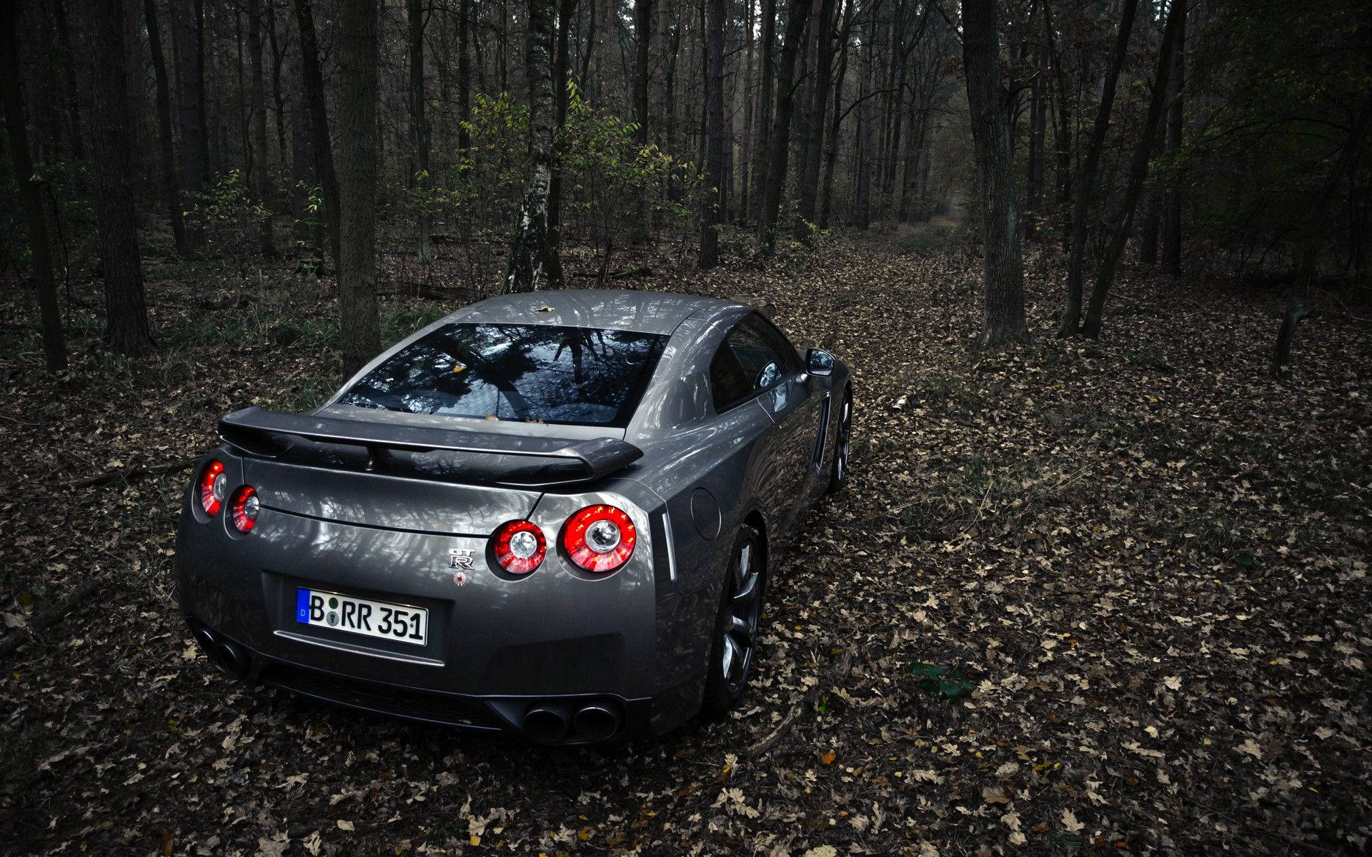 Nissan 桌布 壁紙 — 一輛時尚的灰色跑車，紅色尾燈發光，停在鋪滿落葉的森林小徑上，光線昏暗，氣氛陰沉。