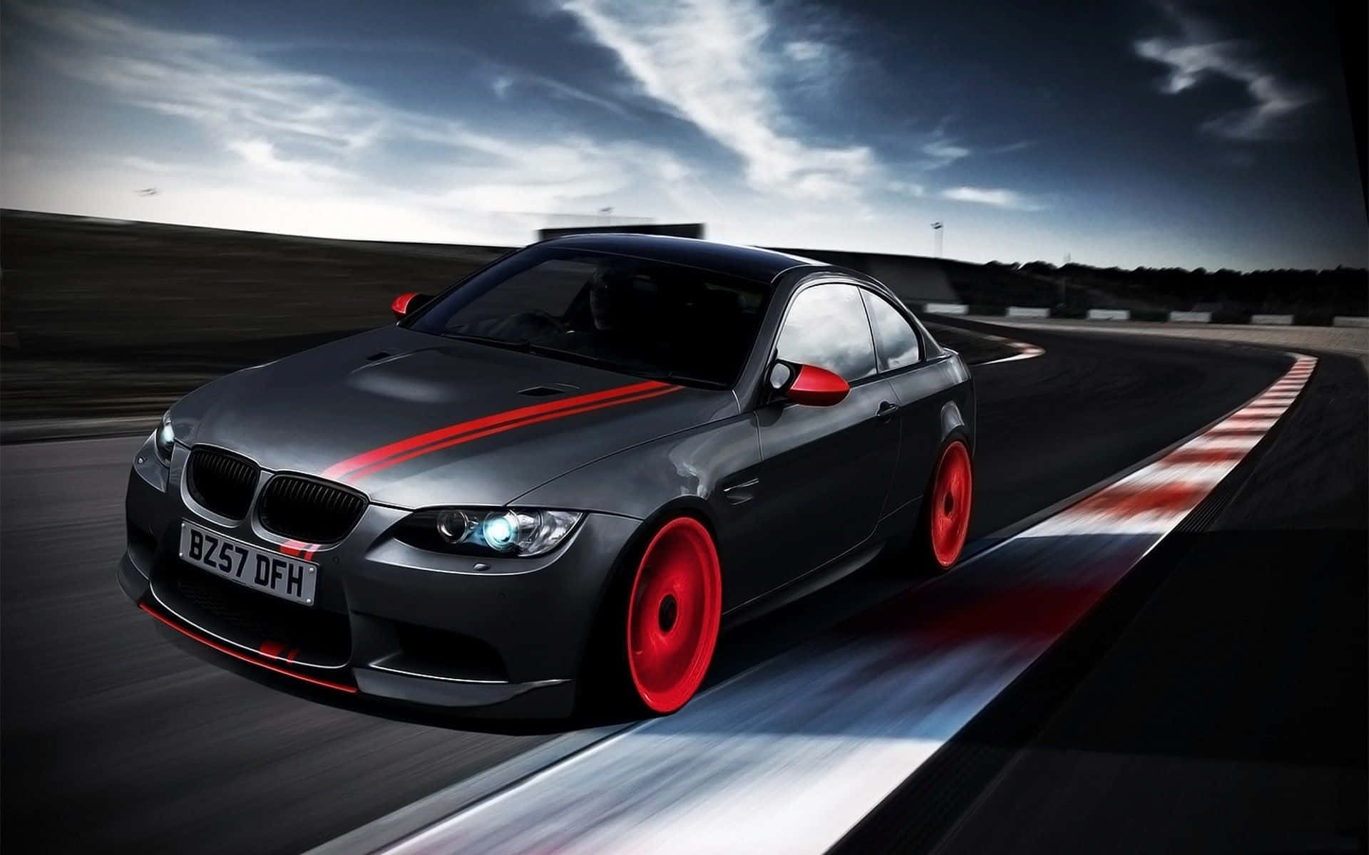Cool Bmw 桌布 壁紙 — 一輛時尚的灰色跑車帶有醒目的紅色點綴，在戲劇性的多雲天空下沿著蜿蜒的賽道疾馳。