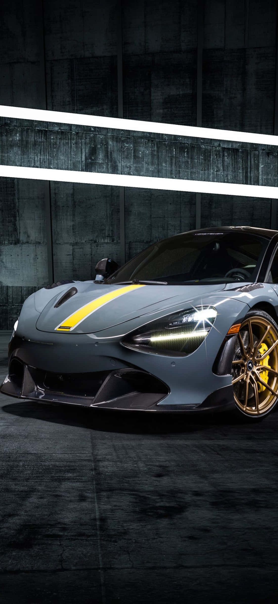 iPhone Xs Mclaren 720s 桌布 壁紙 — 一輛時尚的灰色跑車帶有黃色點綴和金色輪圈，在明亮的白色燈光下展示，背景是深色混凝土牆。