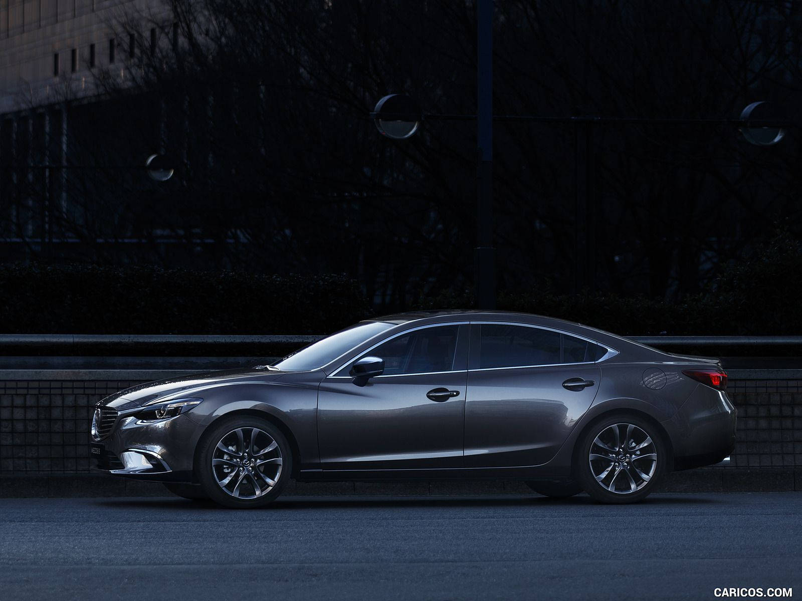 Mazda 桌布 壁紙 — 一輛時尚的灰色轎車停泊在黃昏時分燈光昏暗的城市街道上，背景有光禿的樹木和現代建築。