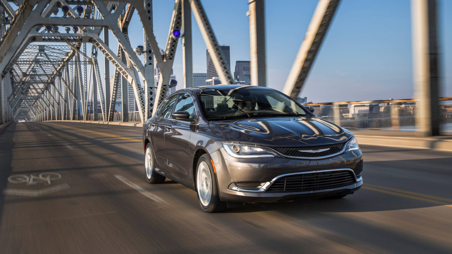 Chrysler 桌布 壁紙 — 一輛時尚的灰色轎車在晴朗的藍天下駛過鋼桁架橋，背景是城市天際線。