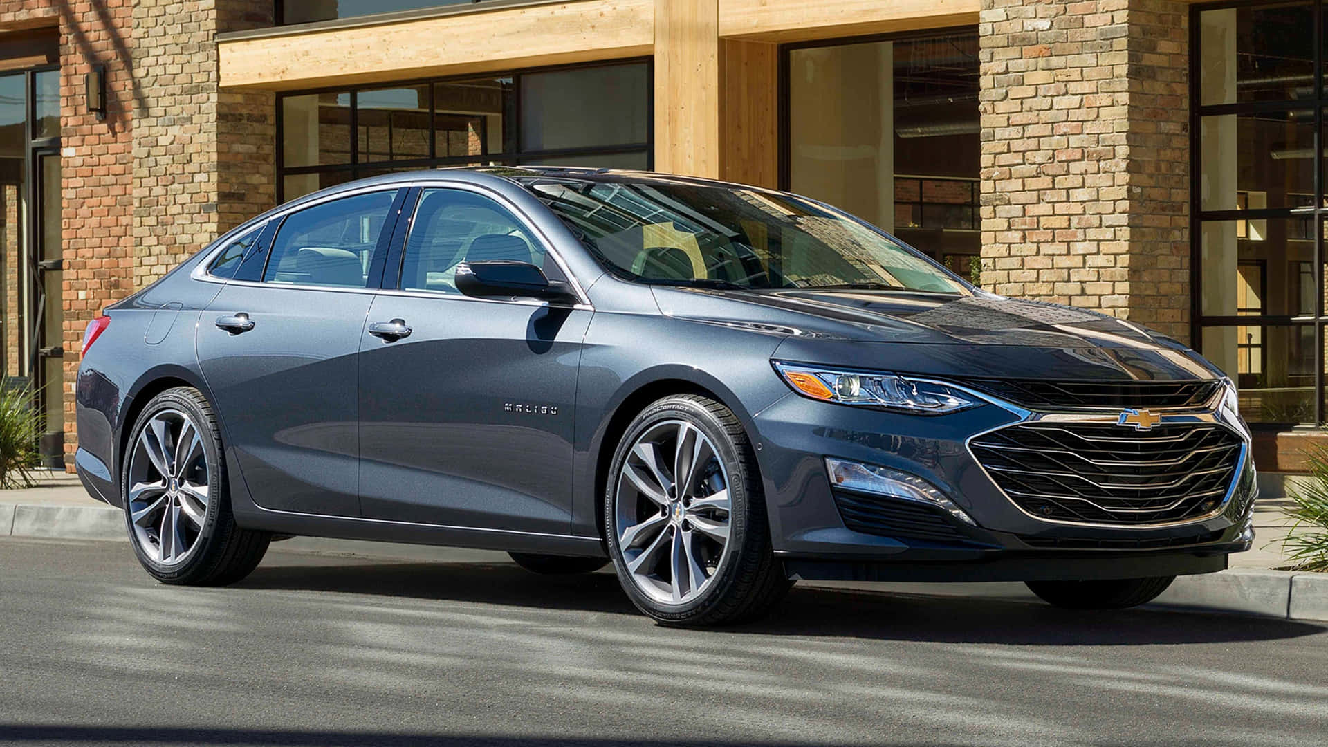 Chevy Malibu 桌布 壁紙 — 一輛時尚的灰色雪佛蘭 Malibu 轎車停放在陽光明媚的街道上，旁邊是磚木建築。
