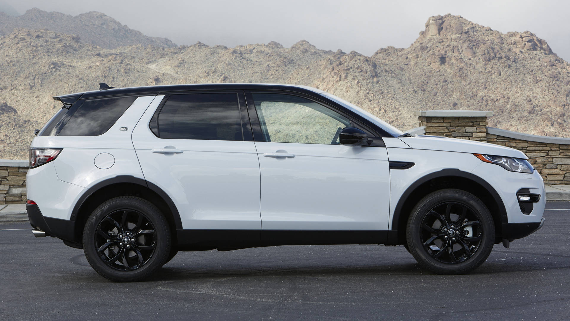 land rover iPhone 桌布 壁紙 — 一輛時尚的白色 Land Rover Discovery Sport 帶有黑色飾邊，停在瀝青路面上，背景是崎嶇的沙漠山脈。