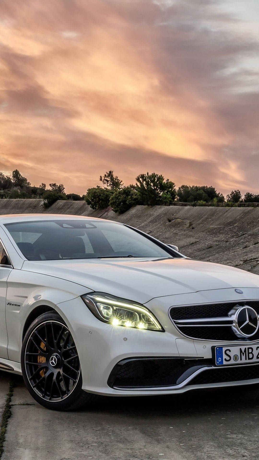 賓士 iPhone 桌布 壁紙 — 一輛時尚的白色 Mercedes-AMG 跑車在充滿活力的日落天空下閃耀，天空中有橙色和粉紅色的雲彩。