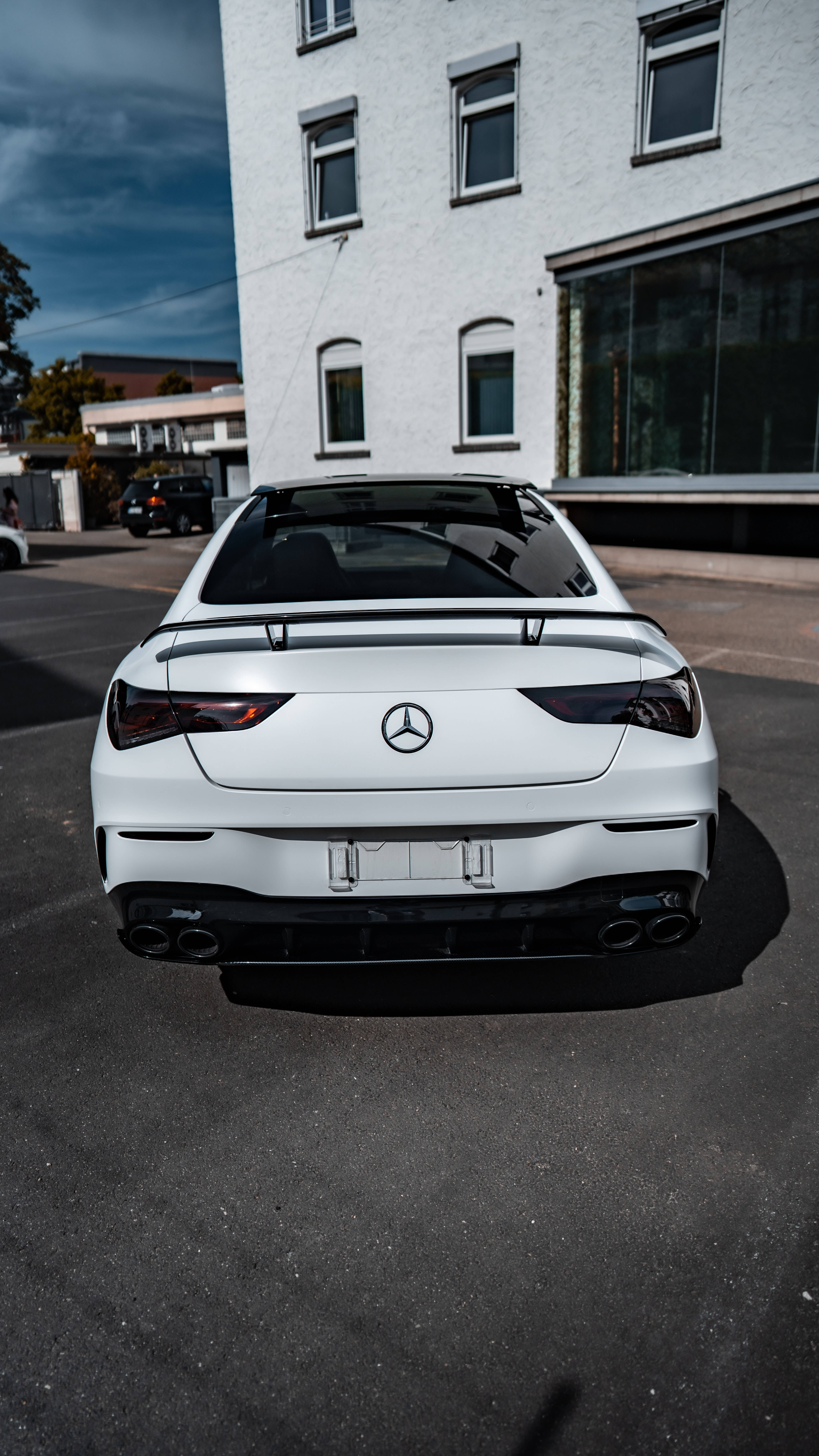 mercedes amg iPhone 桌布 壁紙 — 一輛時尚的白色 Mercedes-AMG 跑車停泊在瀝青路上，背景是現代建築，天空佈滿戲劇性的雲層。