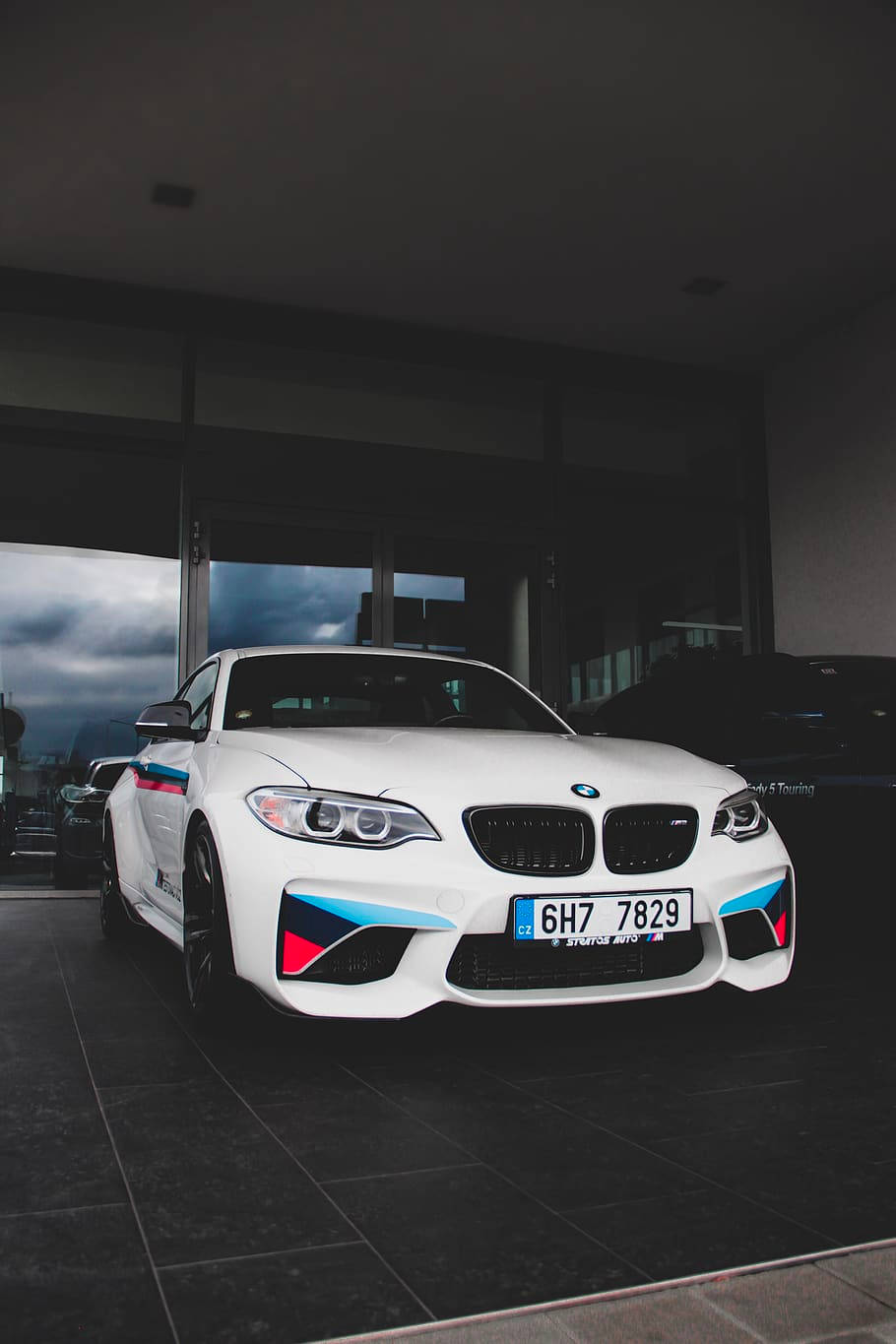 m 桌布 壁紙 — 一輛時尚的白色BMW M2跑車，帶有藍色和紅色點綴，停放在室內深色瓷磚地板上，透過玻璃門可以看到戲劇性的多雲天空。