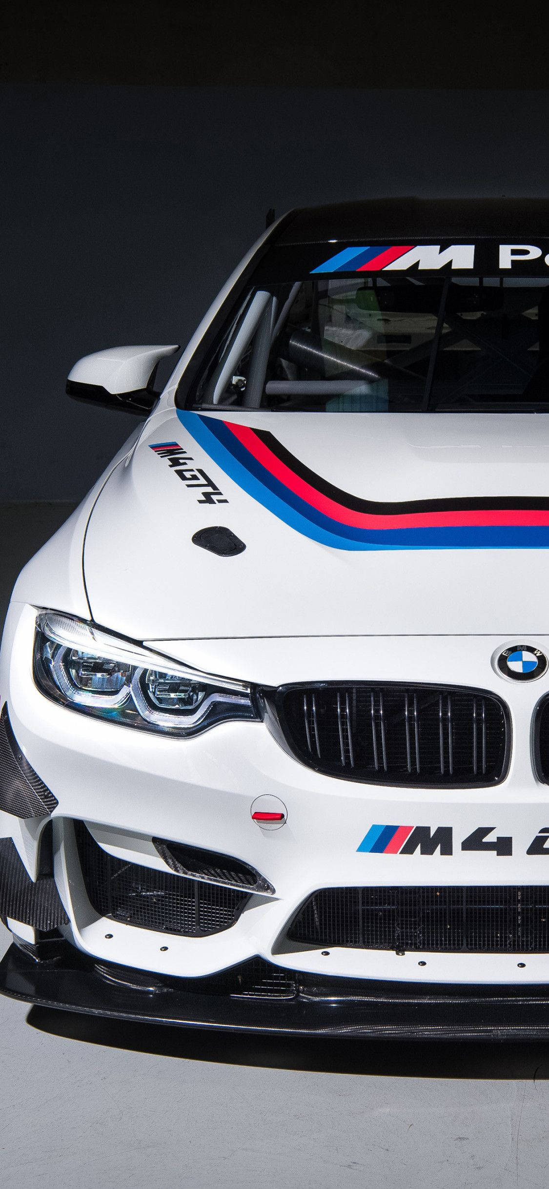 X 桌布 壁紙 — 一輛時尚的白色BMW M4賽車，帶有醒目的藍、紅、黑條紋，在戲劇性的攝影棚拍攝中捕捉到其侵略性的前臉和賽車貼花。