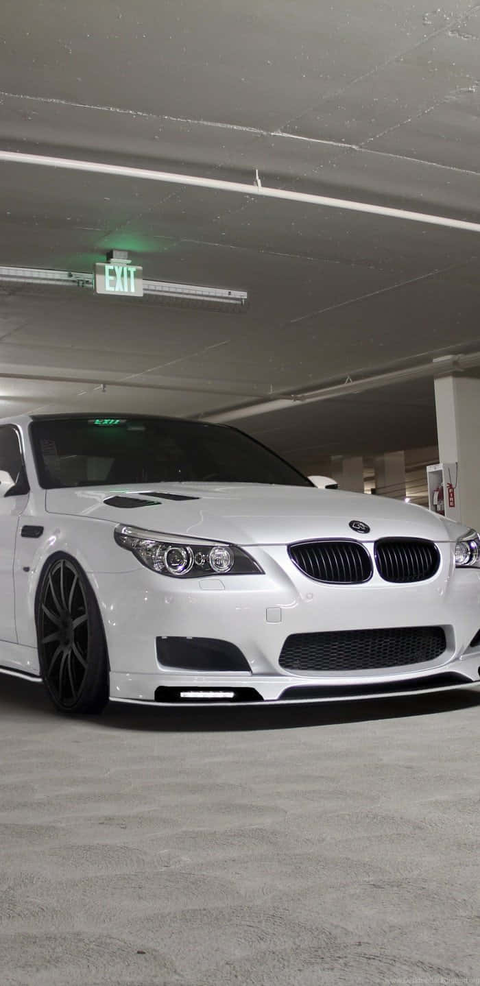 桌布 壁紙 — 一輛時尚的白色BMW M5跑車配黑色輪圈，停泊在昏暗的地下停車場，上方有綠色出口標誌。