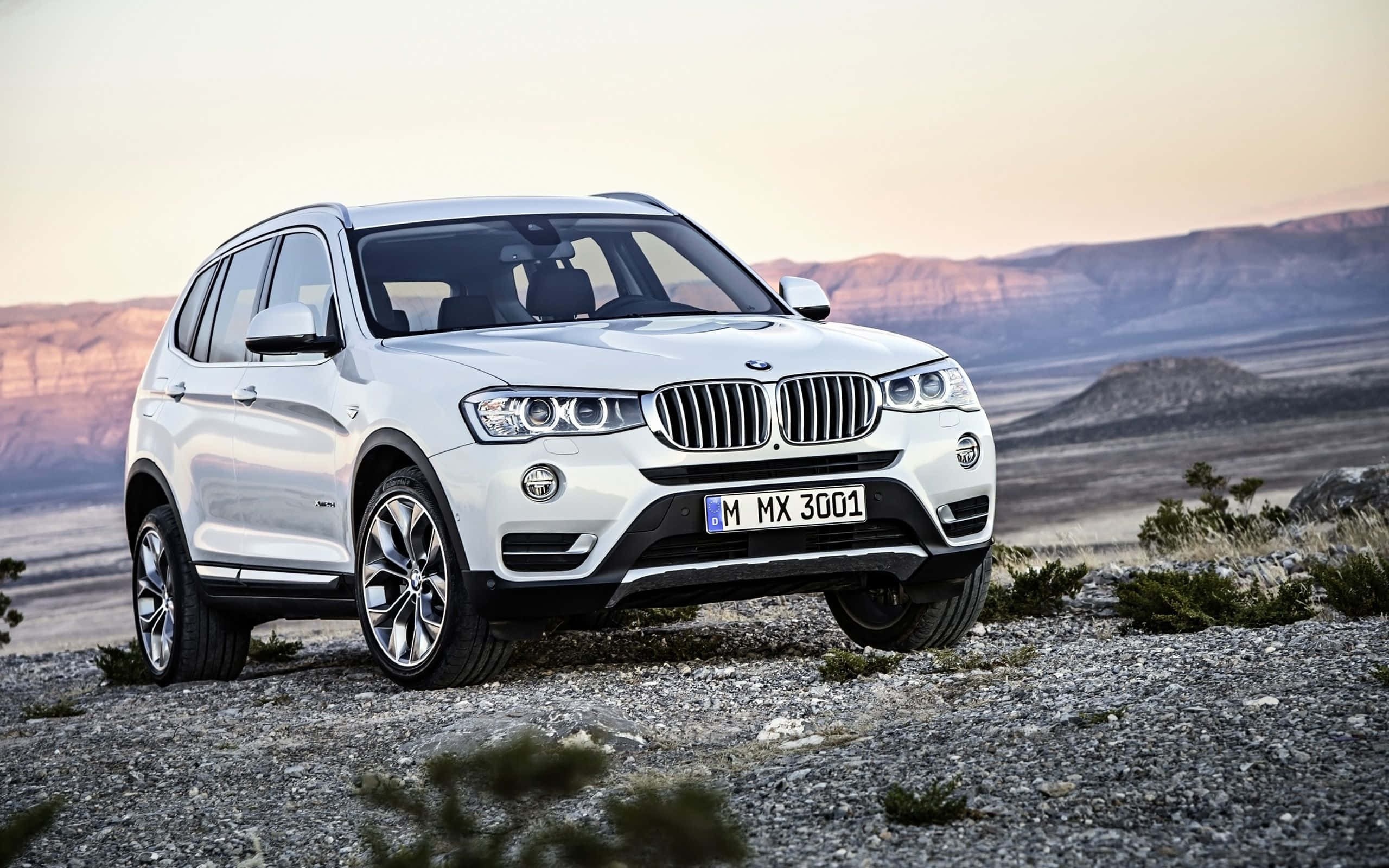 Bmw X3 桌布 壁紙 — 一輛時尚的白色BMW SUV停泊在岩石地形上，背景是沙漠山脈和柔和的日落天空。