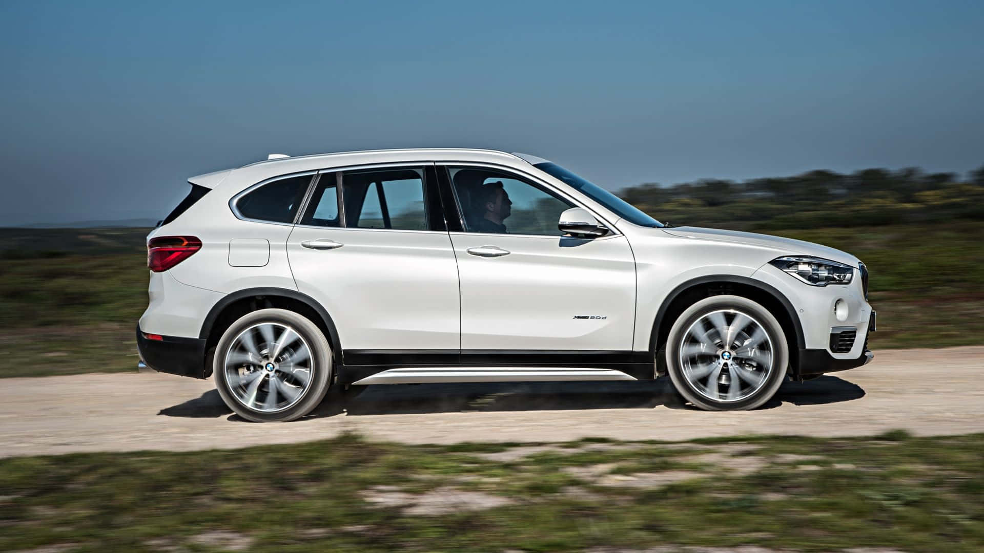 Bmw X1 桌布 壁紙 — 一輛時尚的白色BMW SUV在陽光明媚的泥土路上行駛，背景是模糊的綠意和藍天。