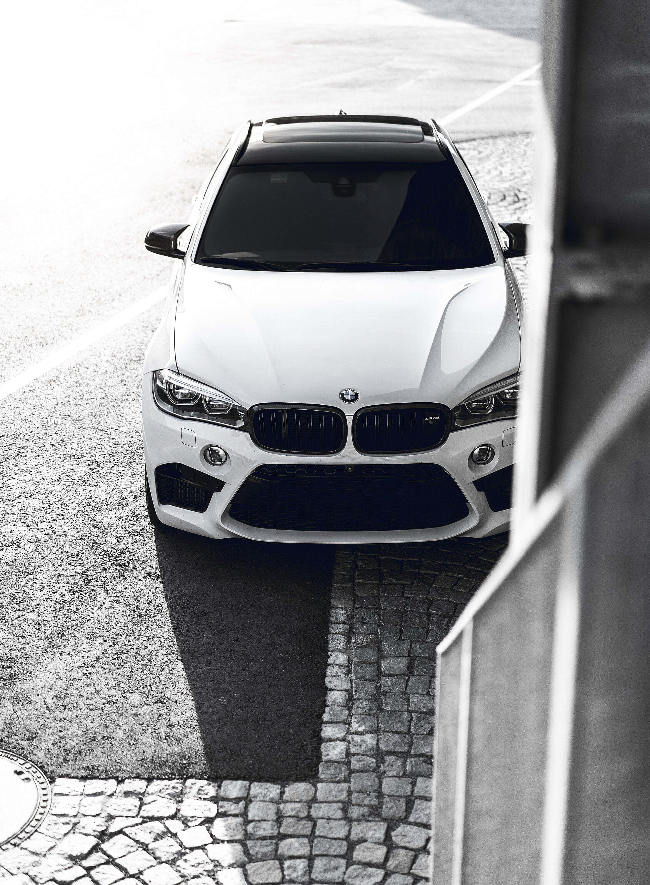 BMW X6 M 桌布 壁紙 — 一輛時尚的白色BMW SUV從上方拍攝，位於鵝卵石街道上，帶有強烈陰影和現代建築框架。