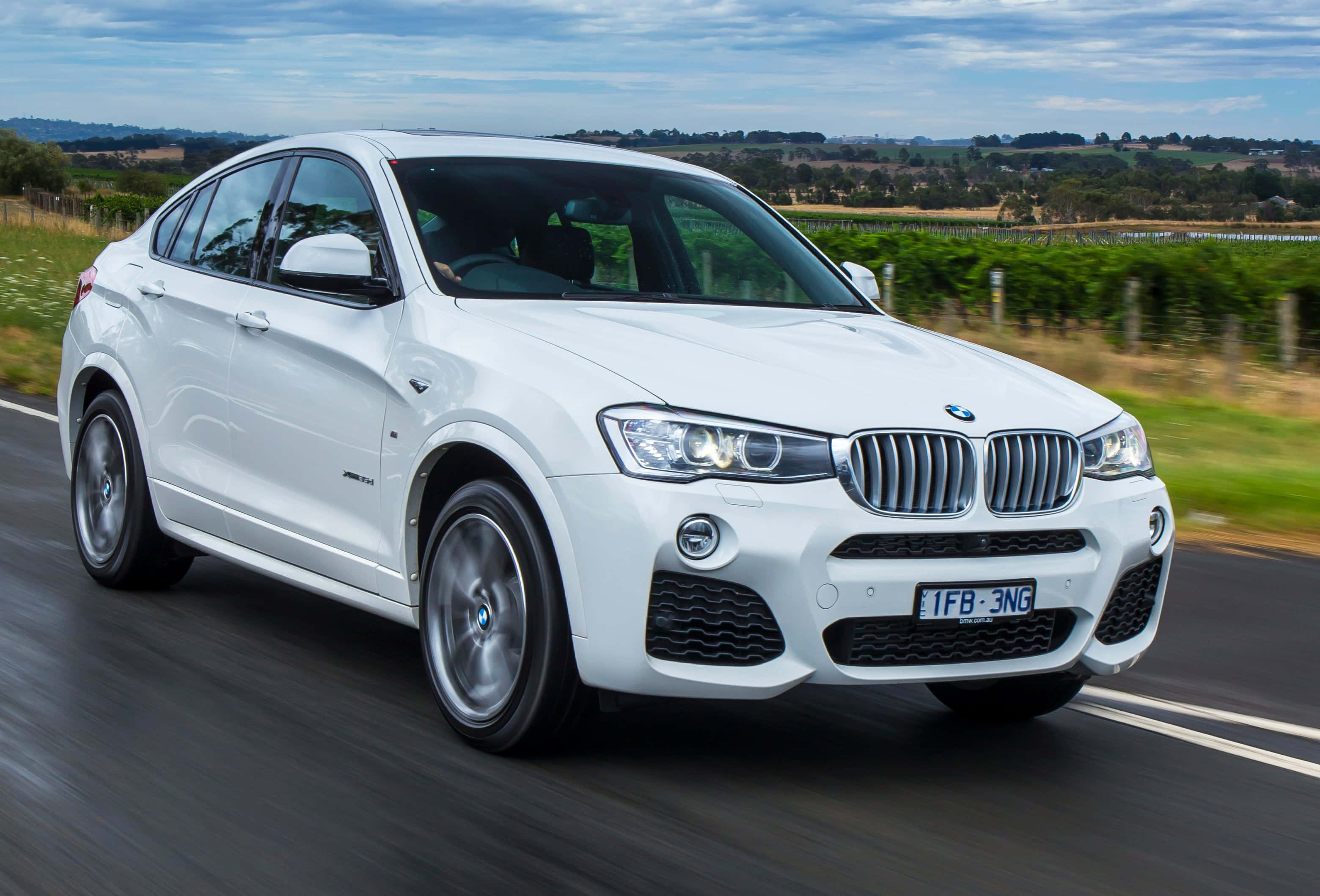Bmw X4 桌布 壁紙 — 一輛時尚的白色BMW X4在鄉村道路上行駛，背景是模糊的綠色田野和葡萄園，天空多雲。