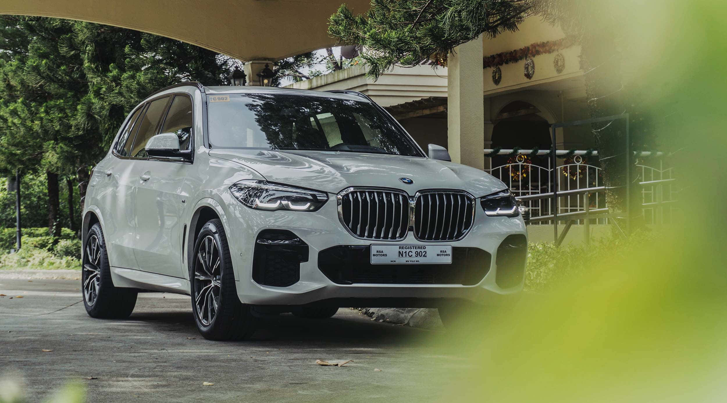 Bmw X5 桌布 壁紙 — 一輛時尚的白色BMW X5 SUV停放在有蓋車道下，周圍環繞著茂密的綠植和一棟經典房屋。
