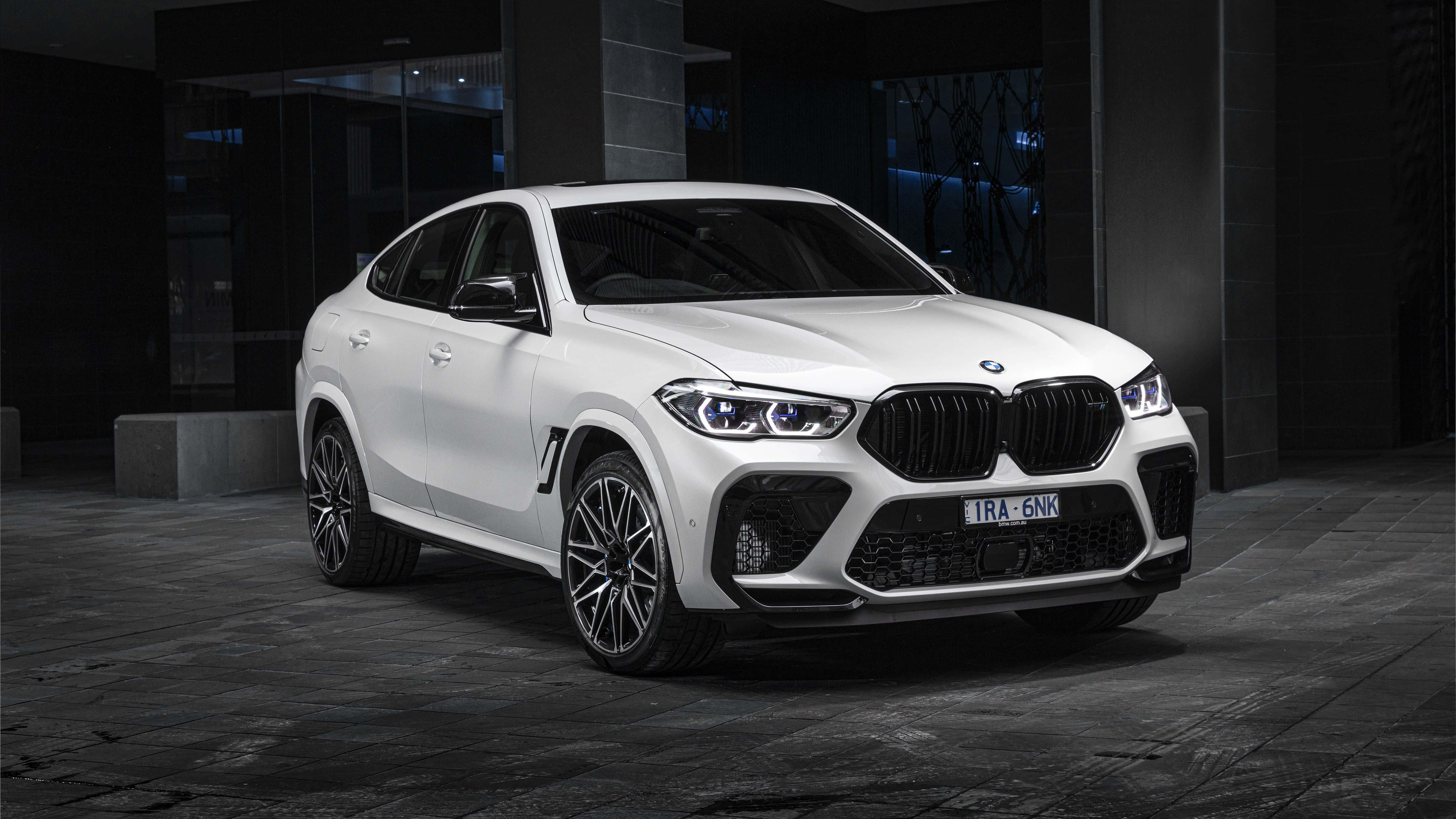 BMW X6 M 桌布 壁紙 — 一輛時尚的白色BMW X6 M停放在現代建築下，車頭燈閃爍，周圍環境昏暗。