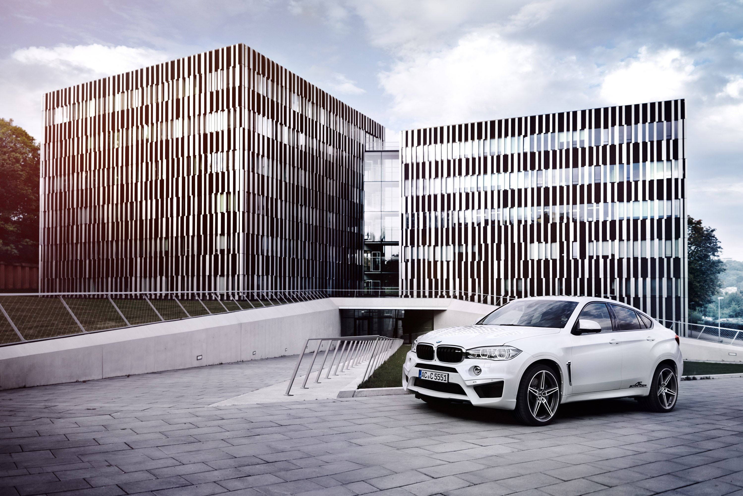 m 桌布 壁紙 — 一輛時尚的白色BMW X6停泊在鋪砌的廣場上，前方是現代建築，其立面帶有垂直板條，天空部分多雲。