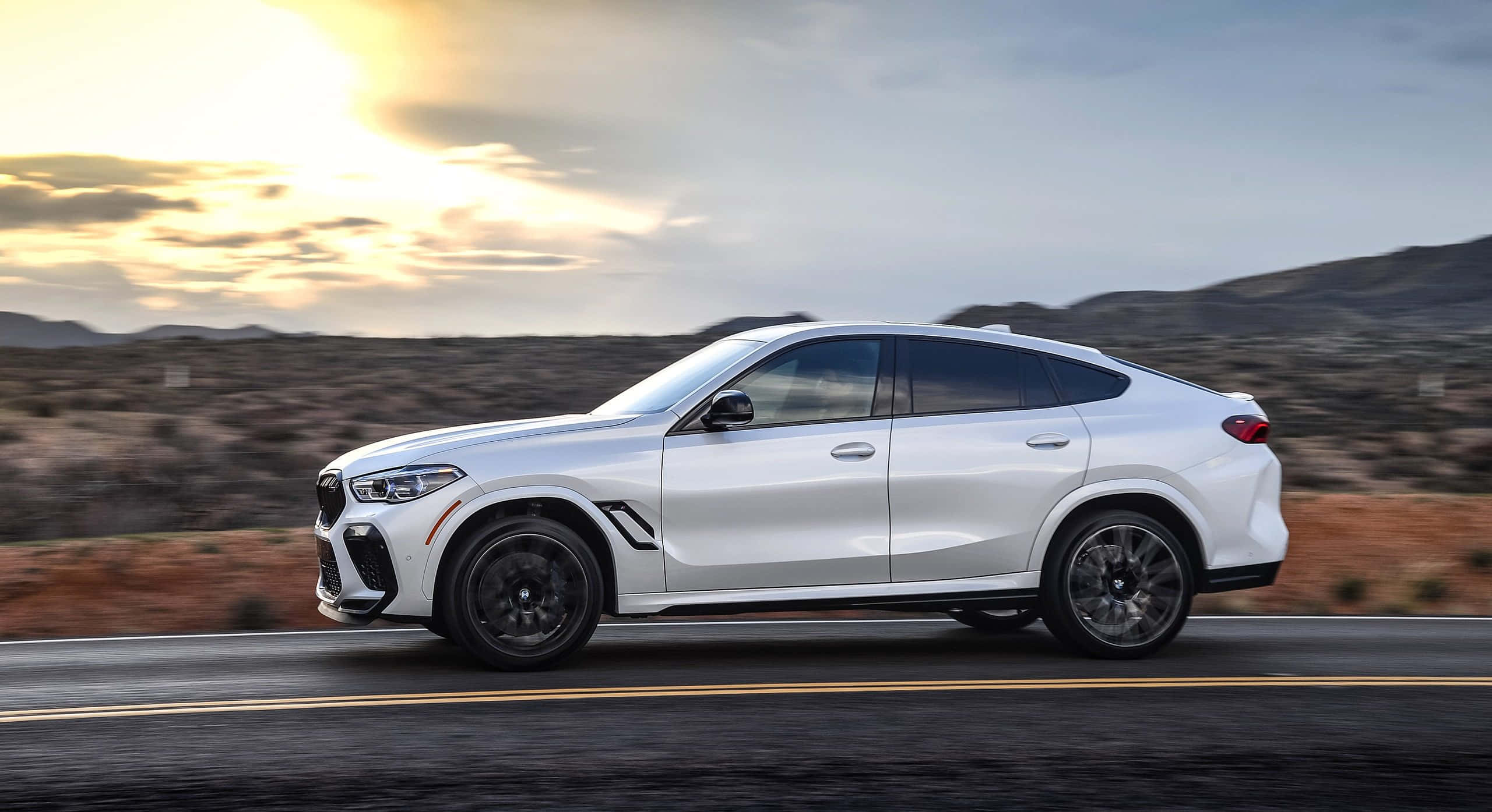 BMW X6 M 桌布 壁紙 — 一輛時尚的白色BMW X6在沙漠公路上行駛，夕陽西下，金色的光芒投下長長的影子。