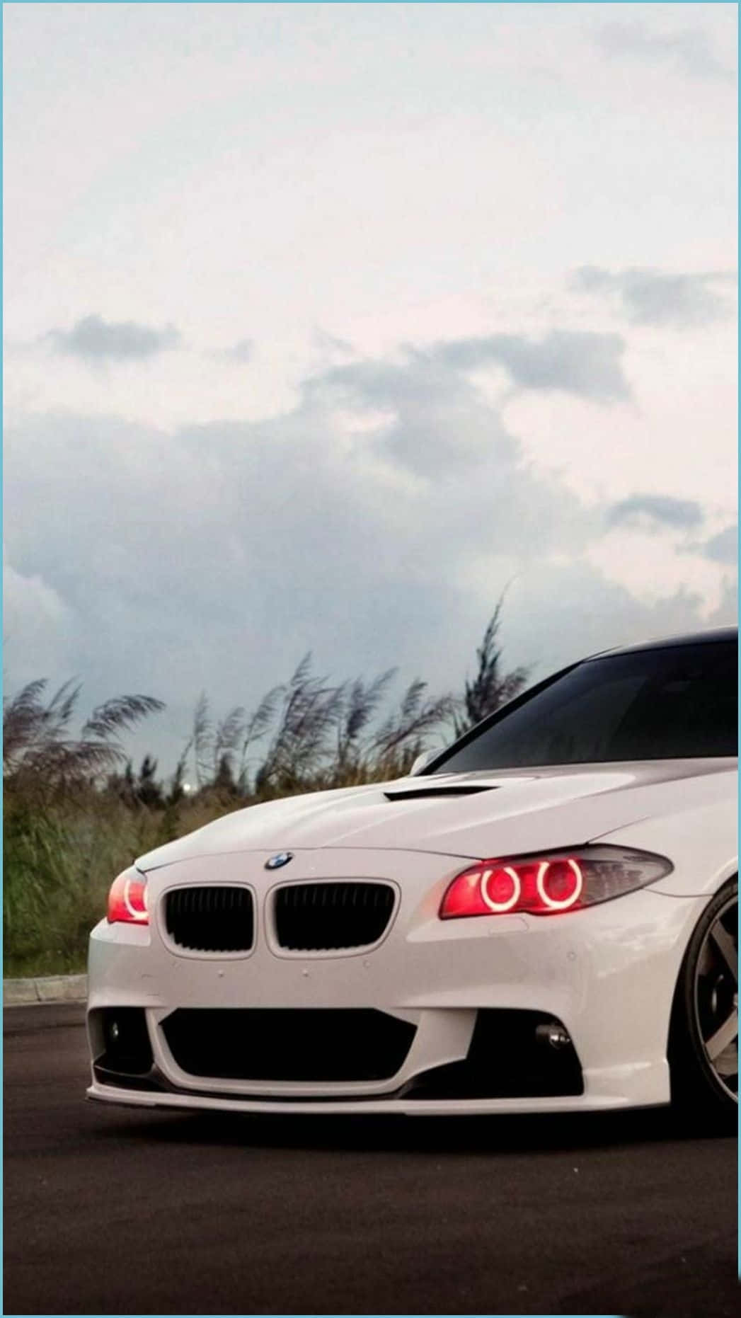 Bmw Android 桌布 壁紙 — 一輛時尚的白色BMW跑車，帶有發光的紅色圓形頭燈，停泊在陰天下高草旁邊的道路上。