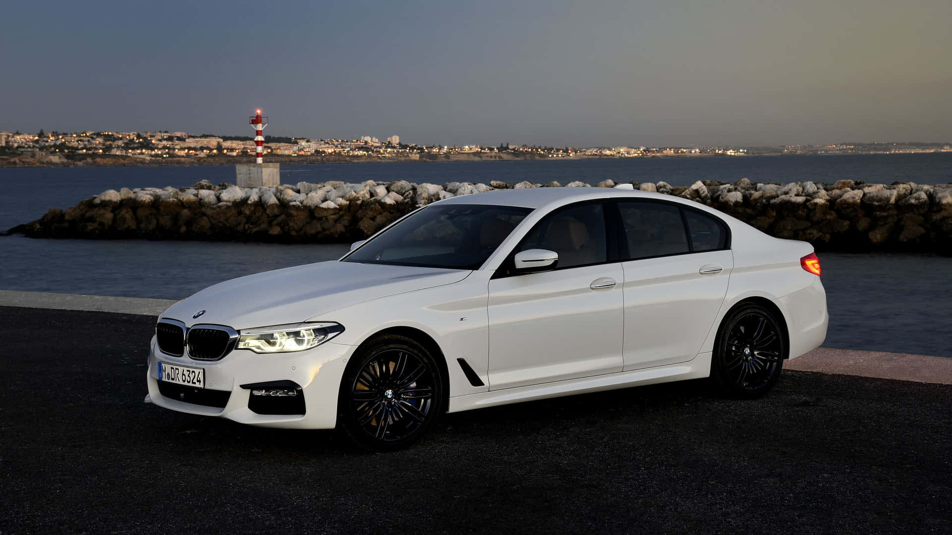 BMW 動圖 桌布 壁紙 — 一輛時尚的白色BMW轎車配黑色輪圈，在黃昏時停靠在海濱，背景有燈塔。