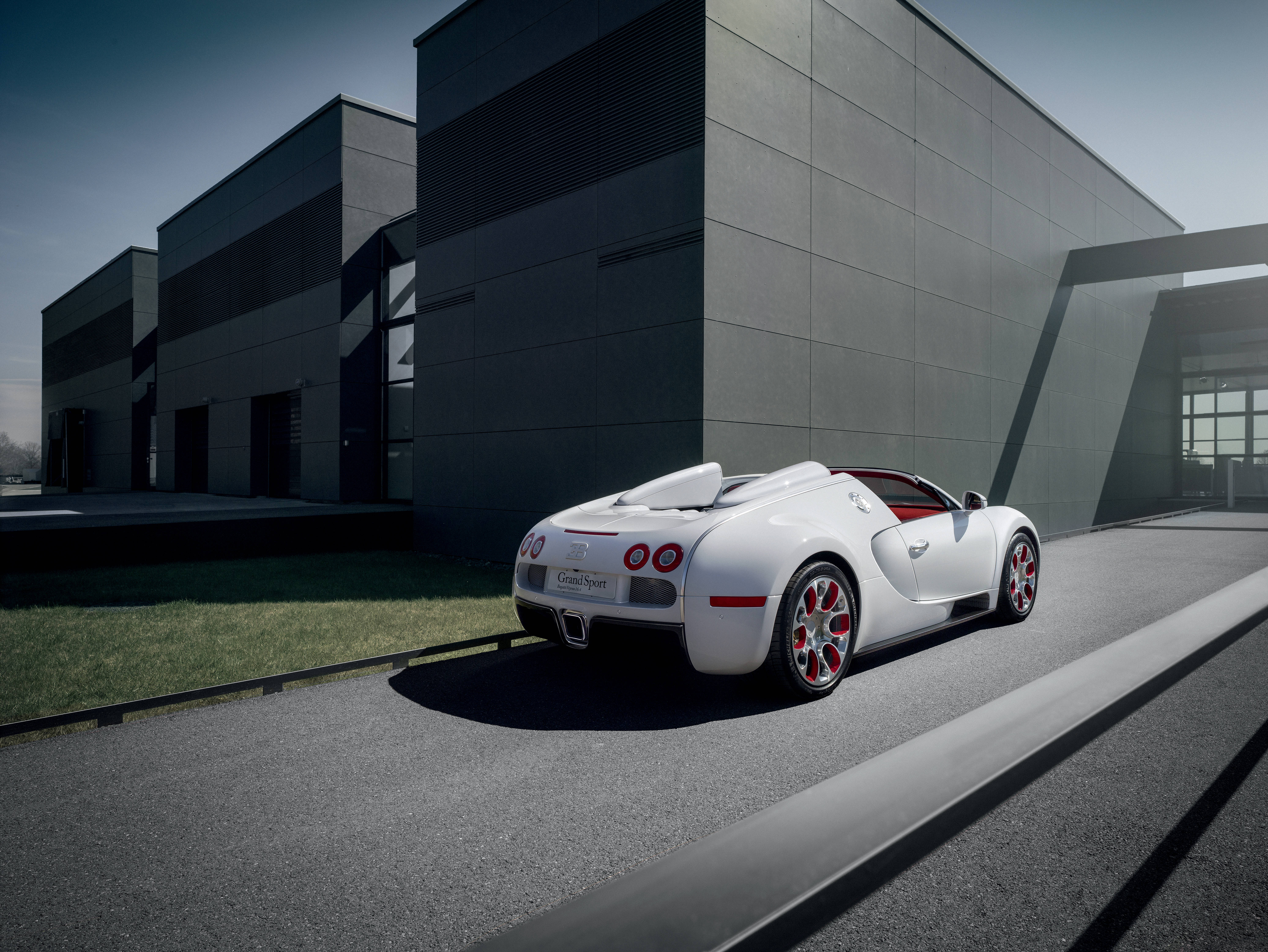 bugatti iPhone 桌布 壁紙 — 一輛時尚的白色Bugatti Veyron Grand Sport敞篷跑車停放在現代灰色路面上，旁邊是簡約工業建築，在晴朗日光下。