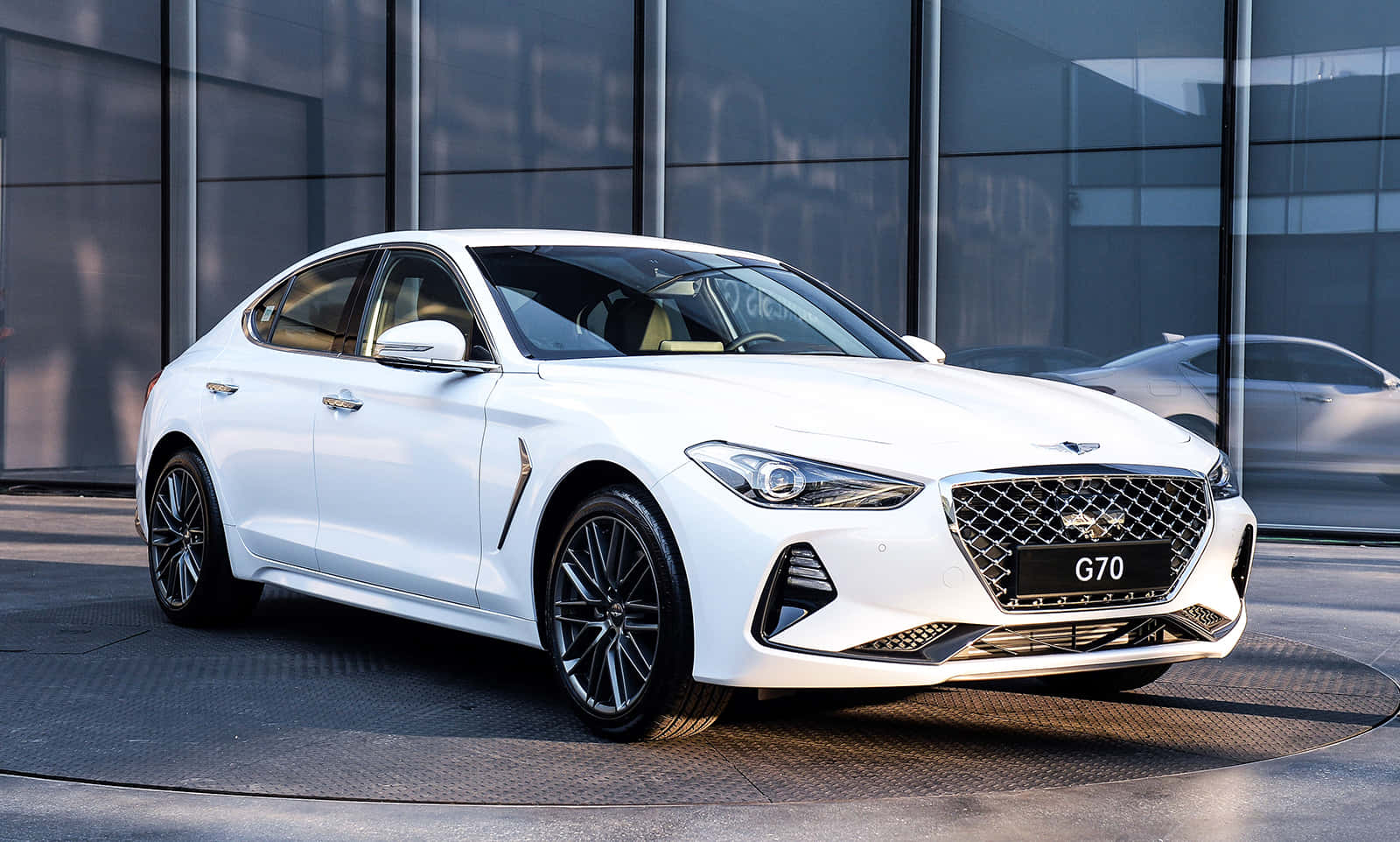 Genesis 桌布 壁紙 — 一輛時尚的白色Genesis G70轎車在明亮的日光下，在現代玻璃建築外牆前閃閃發光。