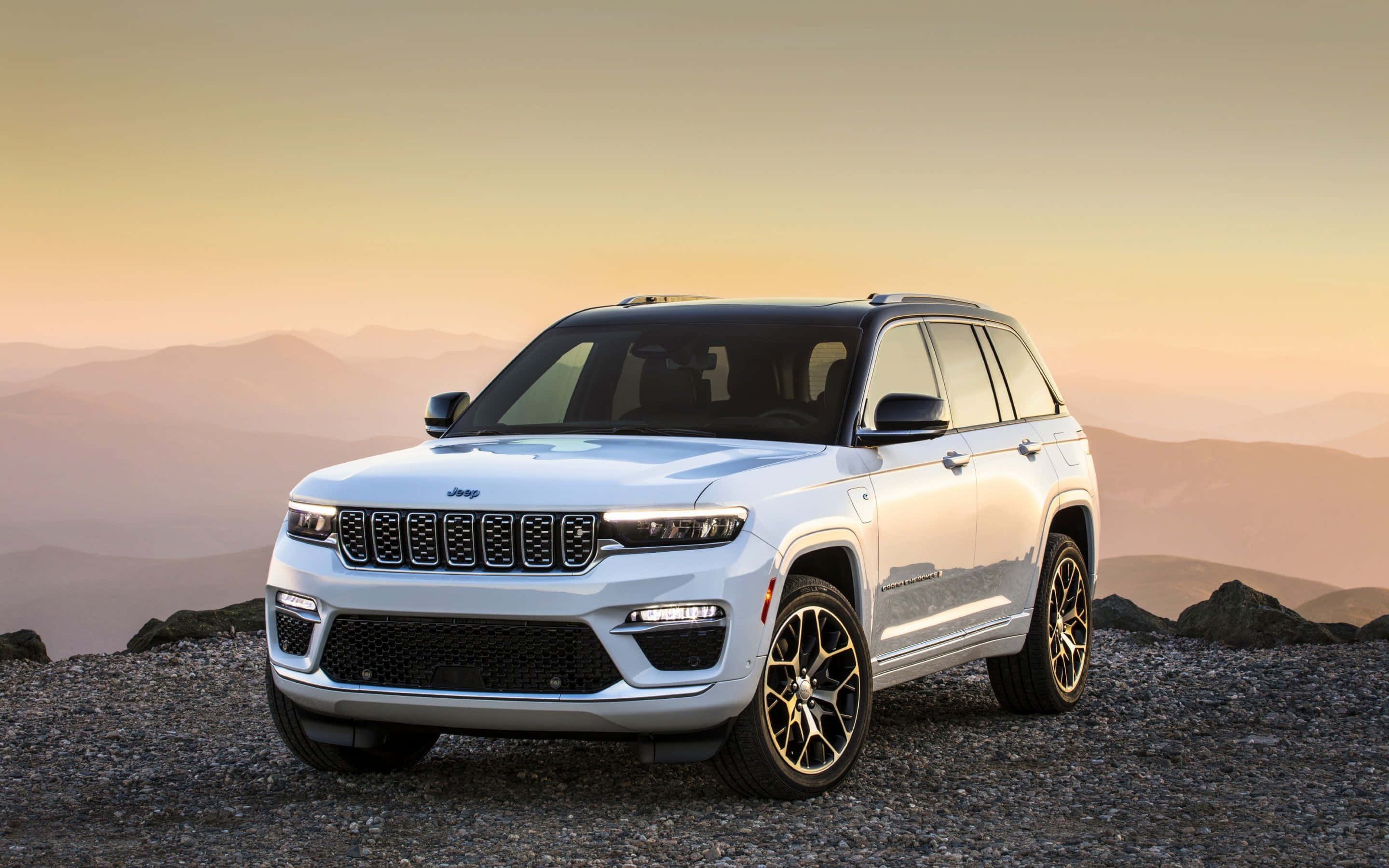 Jeep Wagoneer 桌布 壁紙 — 一輛時尚的白色Jeep Grand Cherokee停泊在日落時分的岩石山脊上，背景是層層疊疊的山脈。