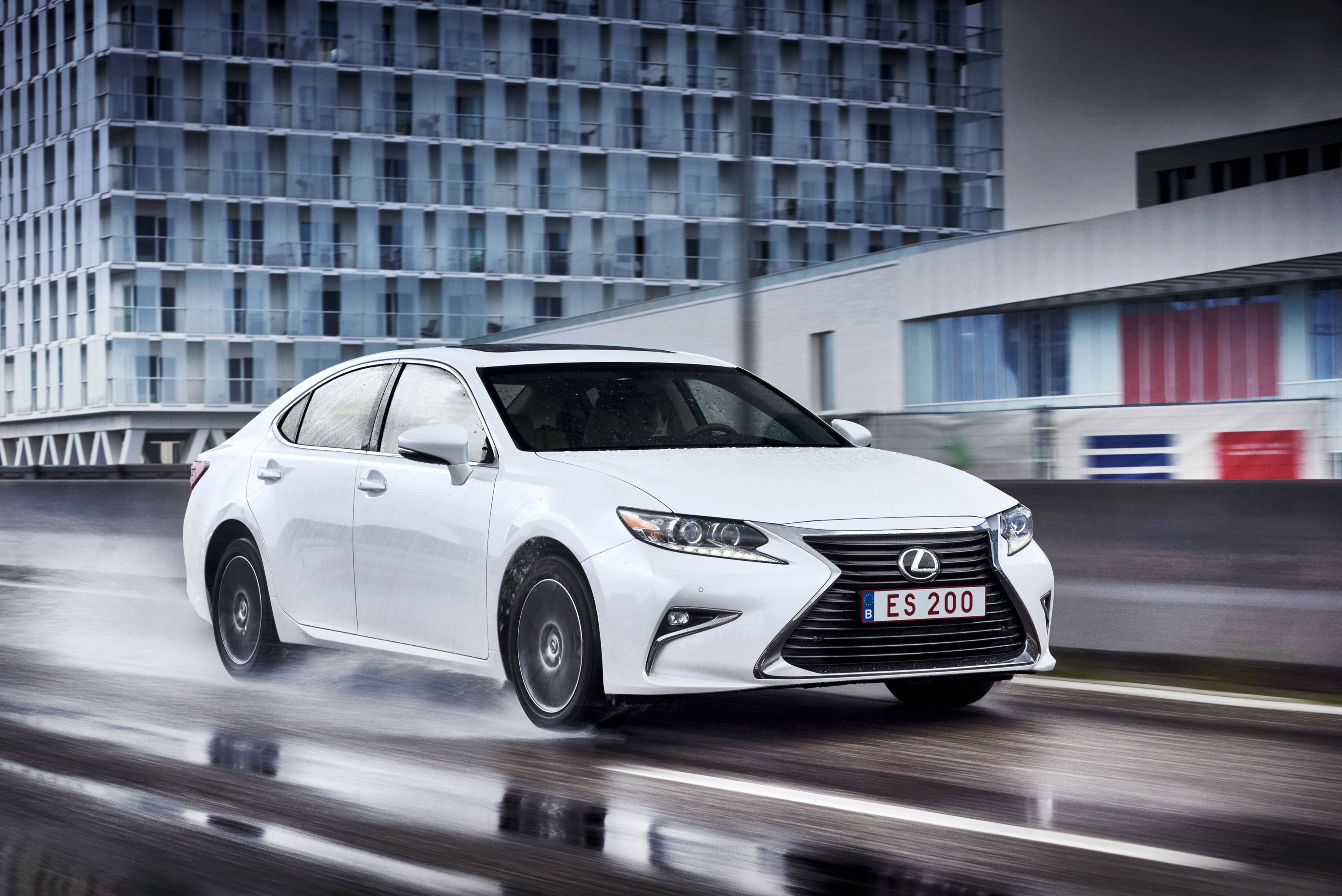 lexus iPhone 桌布 壁紙 — 一輛時尚的白色Lexus ES 200轎車在雨中的城市街道上疾馳，背景有動態模糊和現代玻璃建築。