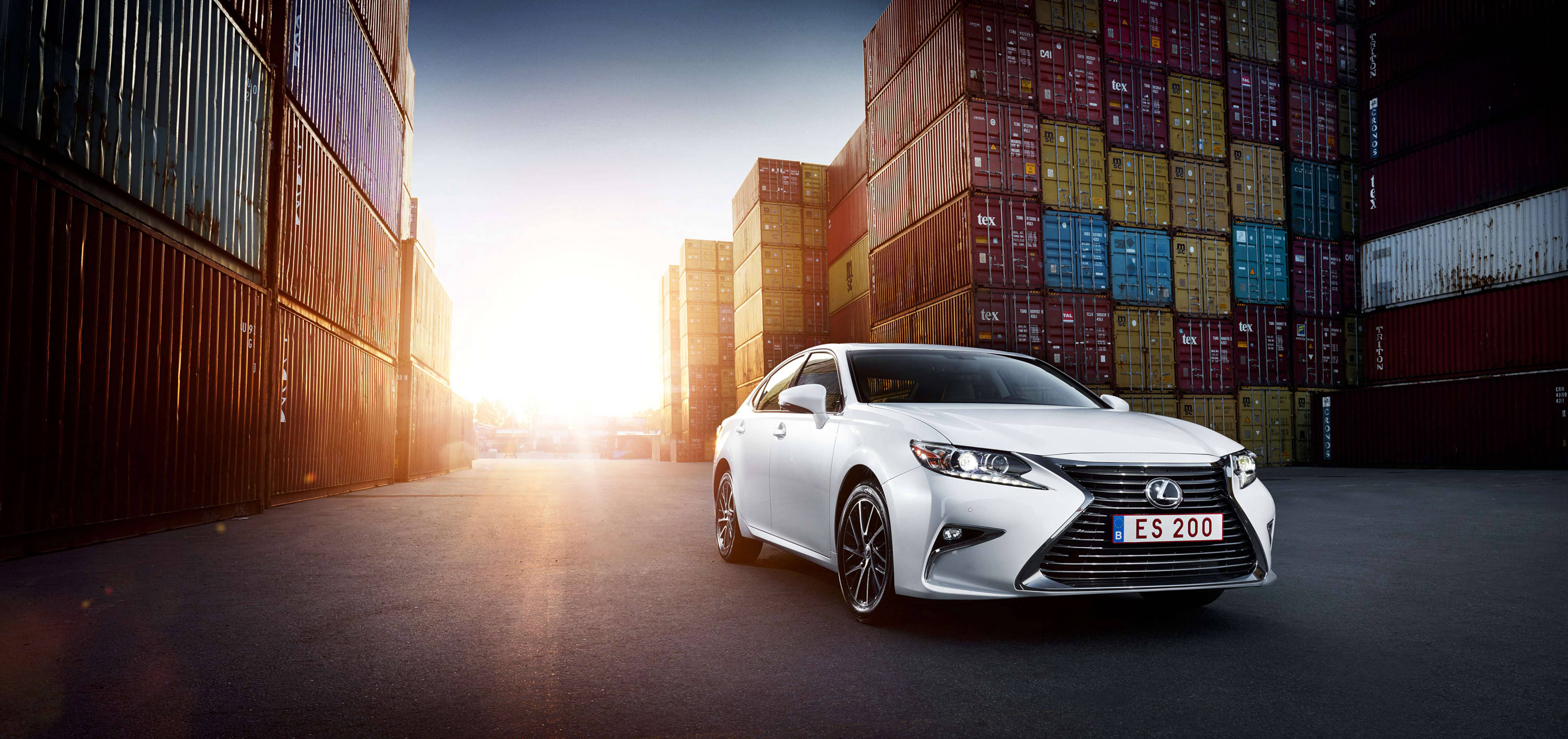 lexus iPhone 桌布 壁紙 — 一輛時尚的白色Lexus ES 200轎車停放在高聳的彩色貨運貨櫃之間，金色的夕陽光芒淹沒了工業小巷。