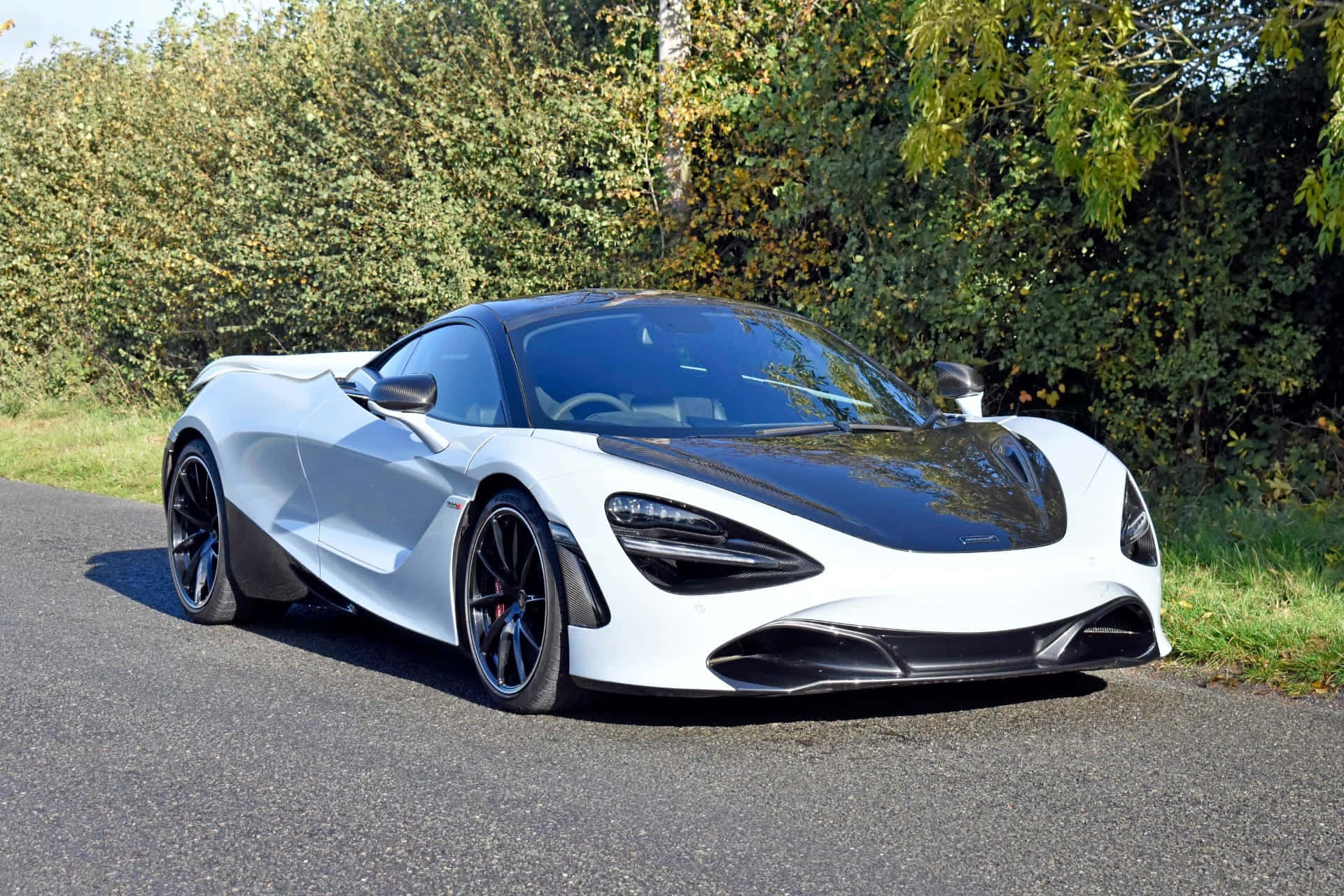 Hood 桌布 壁紙 — 一輛時尚的白色McLaren 720S跑車，帶有黑色飾邊，停在鋪砌的道路旁，旁邊是綠色植物，在明亮的日光下。