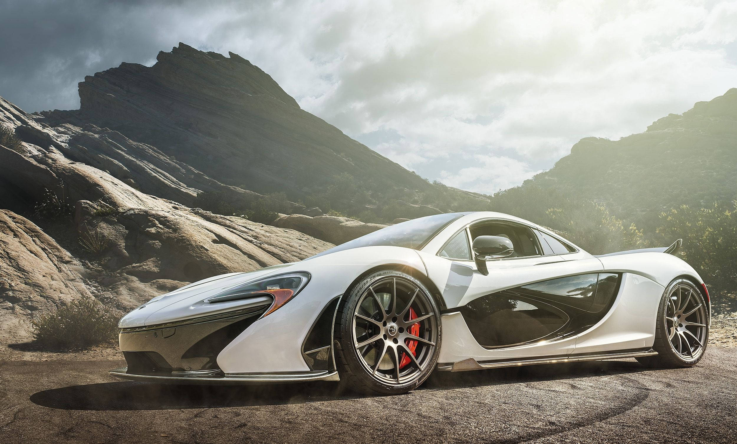 麥拉倫 桌布 壁紙 — 一輛時尚的白色McLaren跑車，帶有紅色制動卡鉗，停泊在一條塵土飛揚的道路上，背景是戲劇性的岩石山脈，天空多雲。
