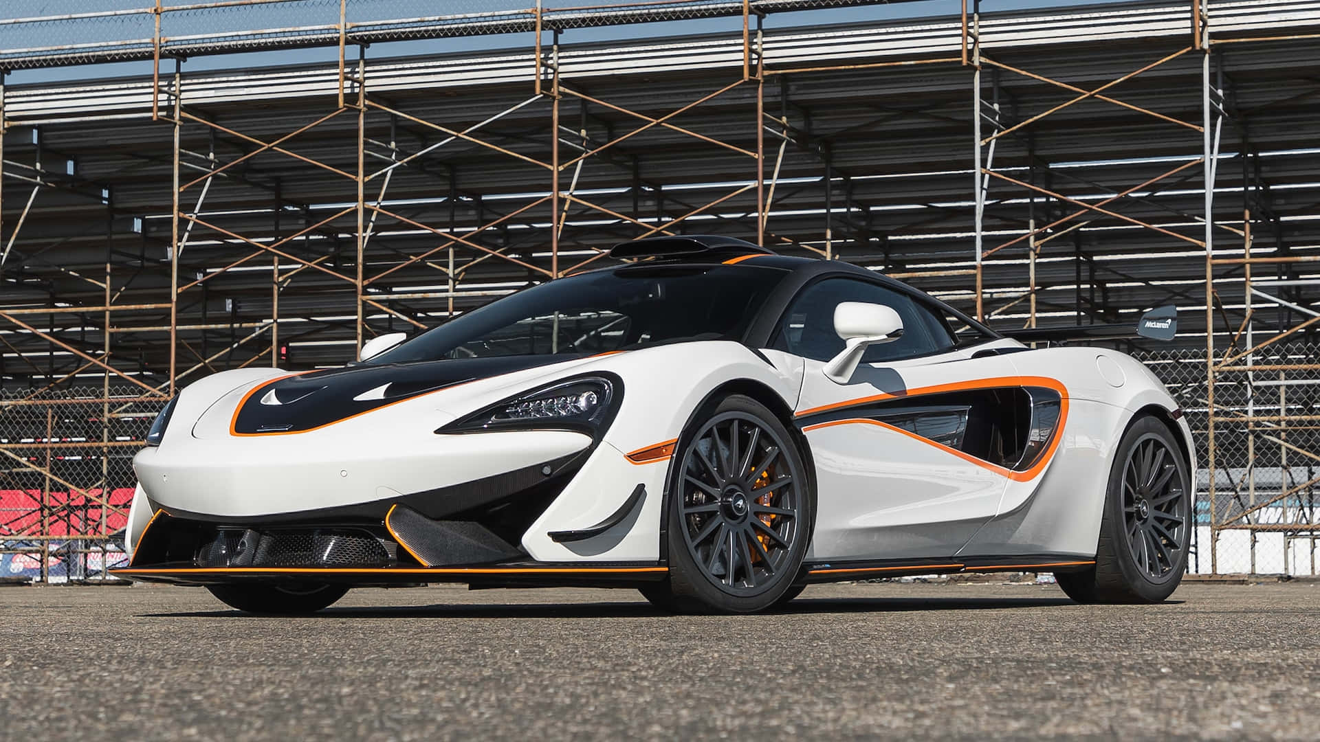 麥拉倫 桌布 壁紙 — 一輛時尚的白色McLaren跑車帶有橙色點綴，停在金屬看台結構下的瀝青路面上。